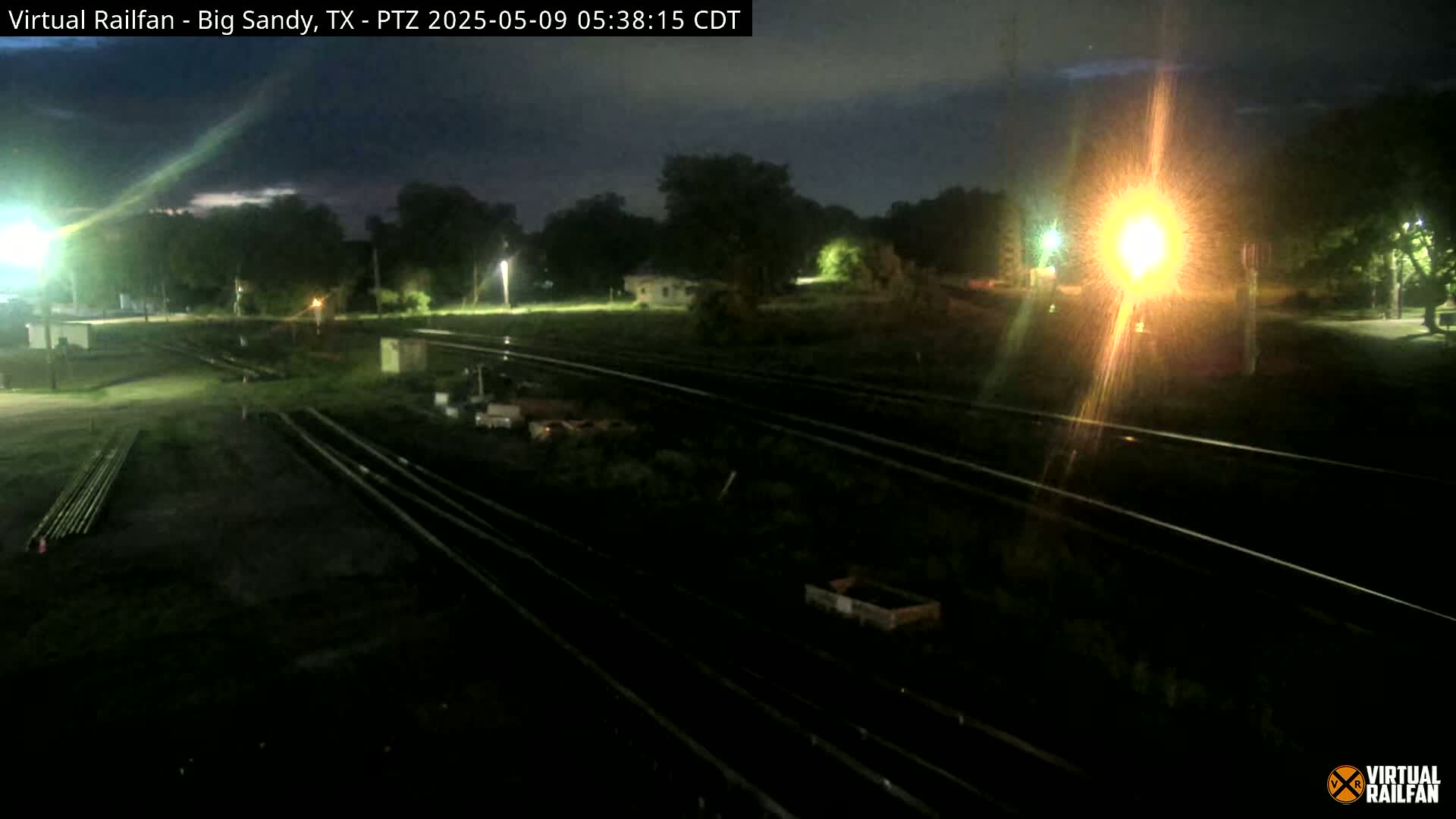 Under a dark, nighttime sky, several sets of train tracks are visible, with some scattered debris and equipment alongside them, near a sparsely lit area with some trees and buildings in the distance.