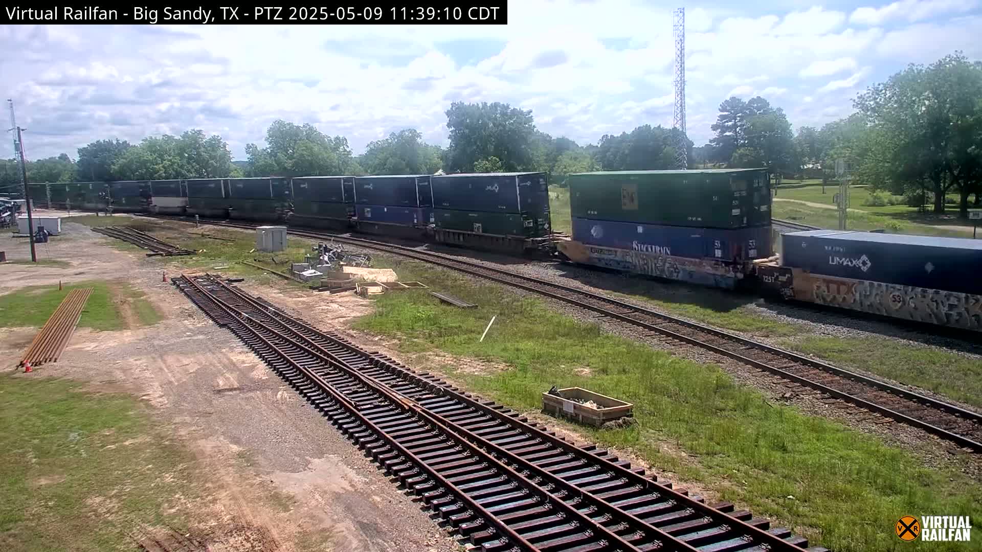 A long freight train carrying multiple shipping containers sits on railroad tracks under a partly cloudy sky, next to a grassy area with some construction materials.