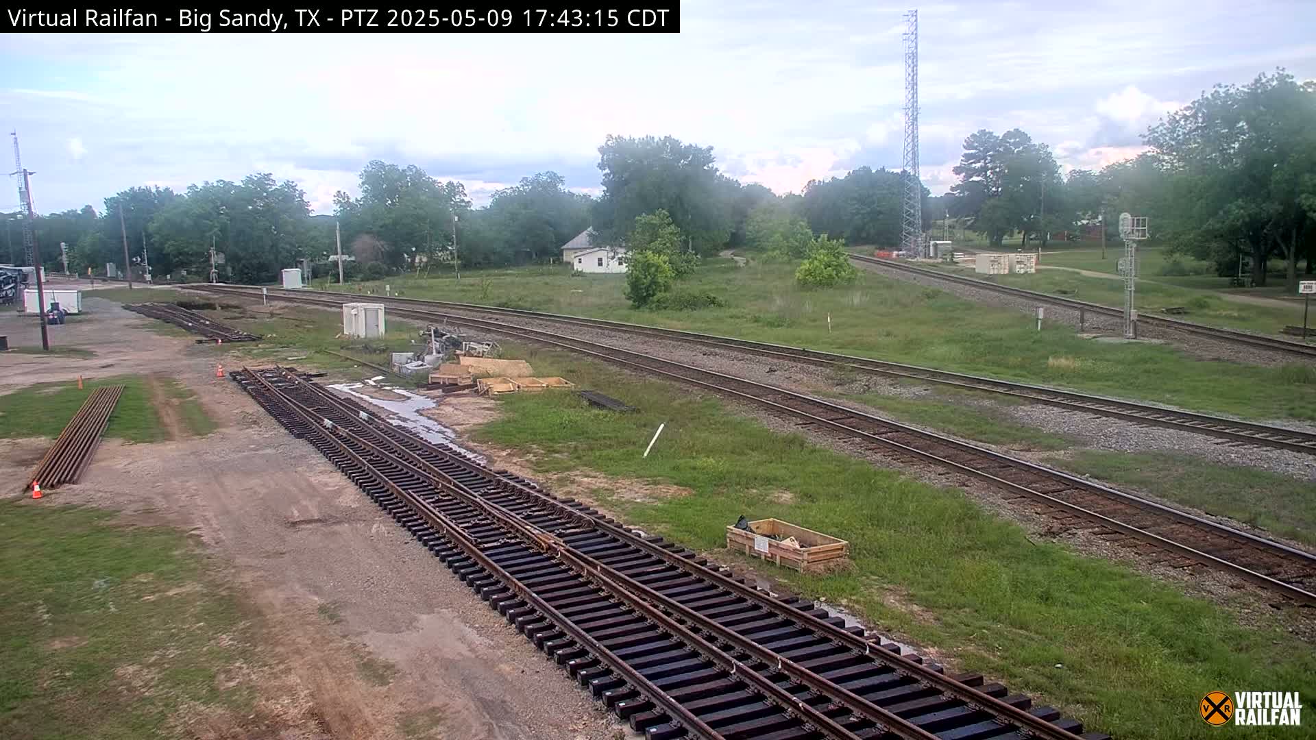 Railroad tracks undergoing construction or maintenance are situated in a grassy area near a small town under an overcast sky.