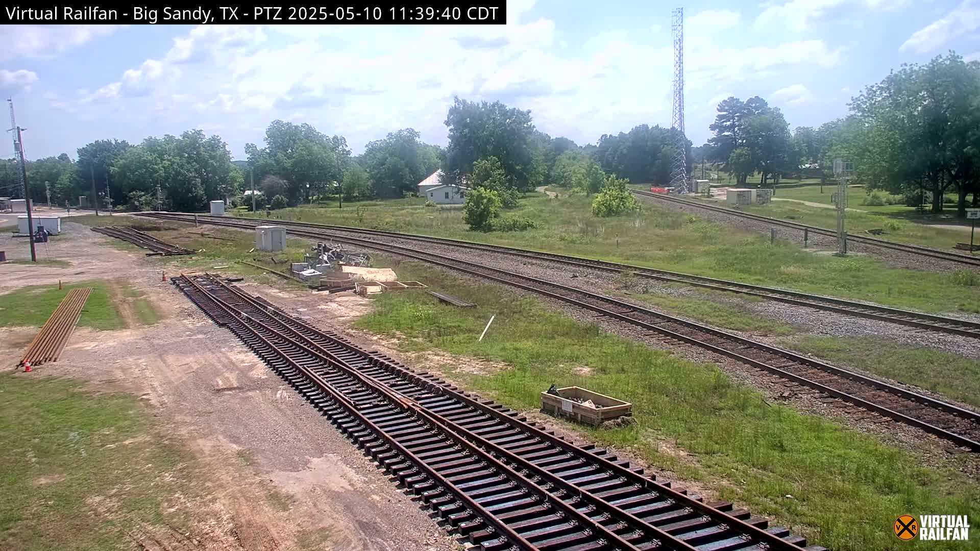 Under a partly cloudy sky, railroad tracks wind through a grassy area with some trees and buildings in the background.