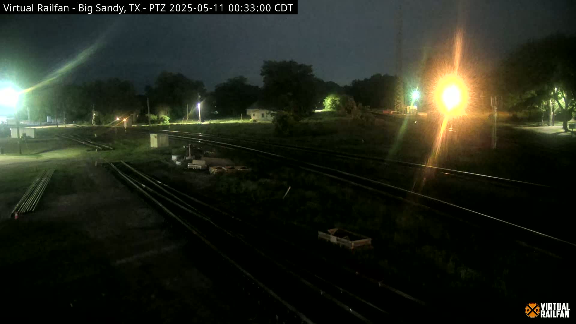 Under a dark night sky, several sets of train tracks wind through a dimly lit area with sparse vegetation, some small structures, and equipment.