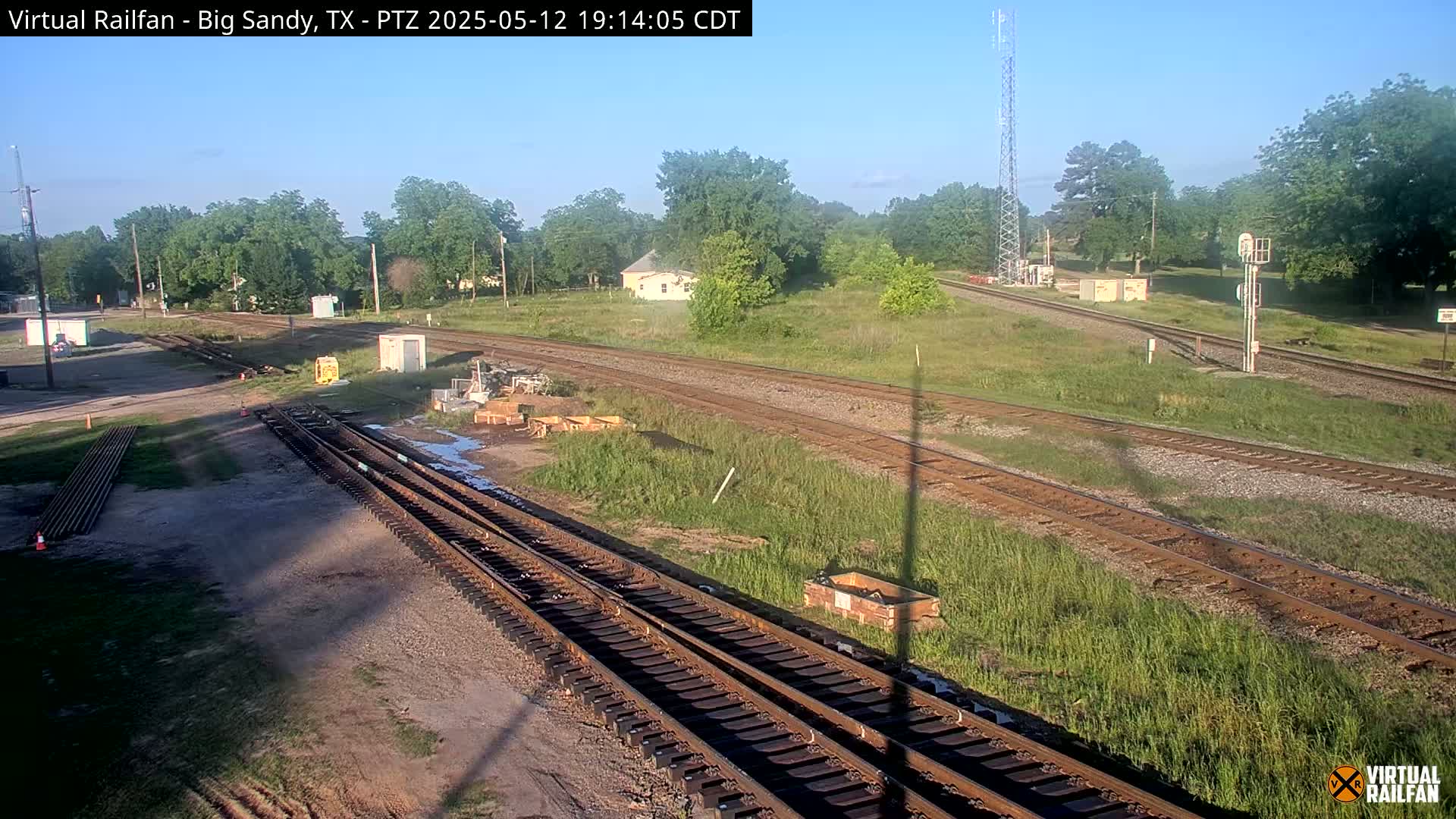 A sunny view of several intersecting railroad tracks surrounded by grassy fields and trees, with some construction materials visible near the tracks.