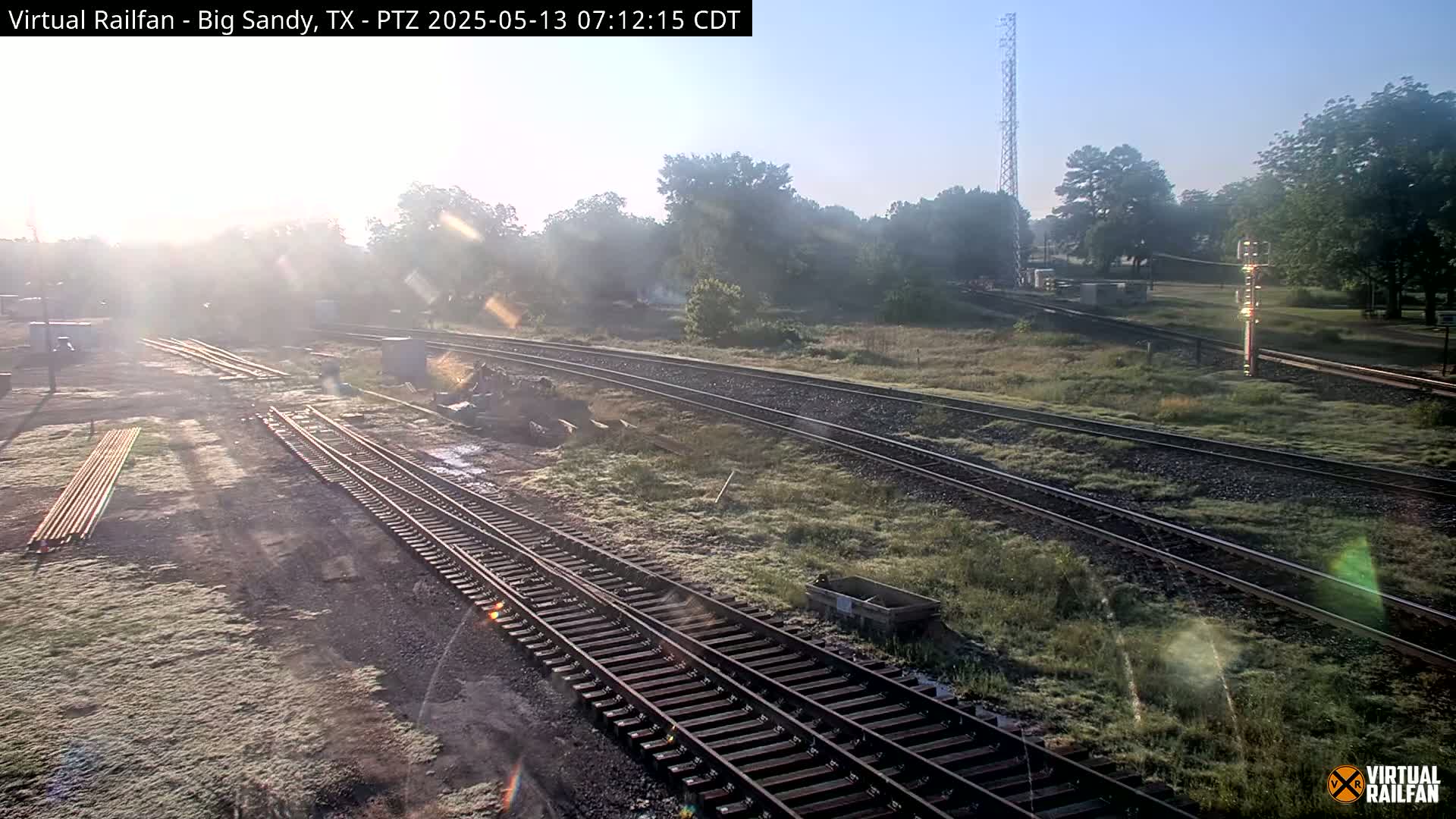 Multiple sets of train tracks wind through a grassy area on a sunny morning with some lens flare.