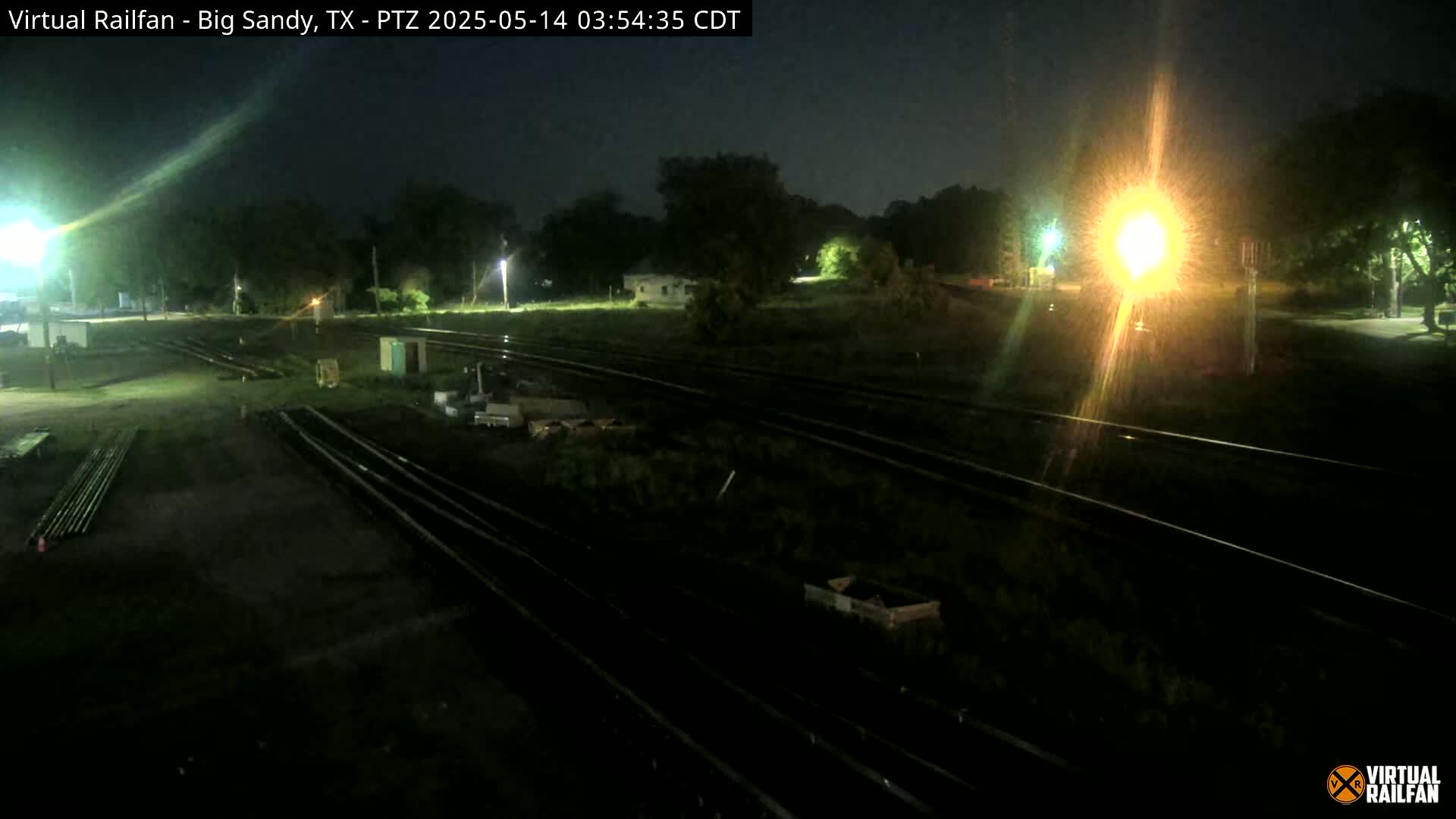 At night, a dark, flat area with train tracks is illuminated by artificial light, showing some equipment and vegetation nearby.