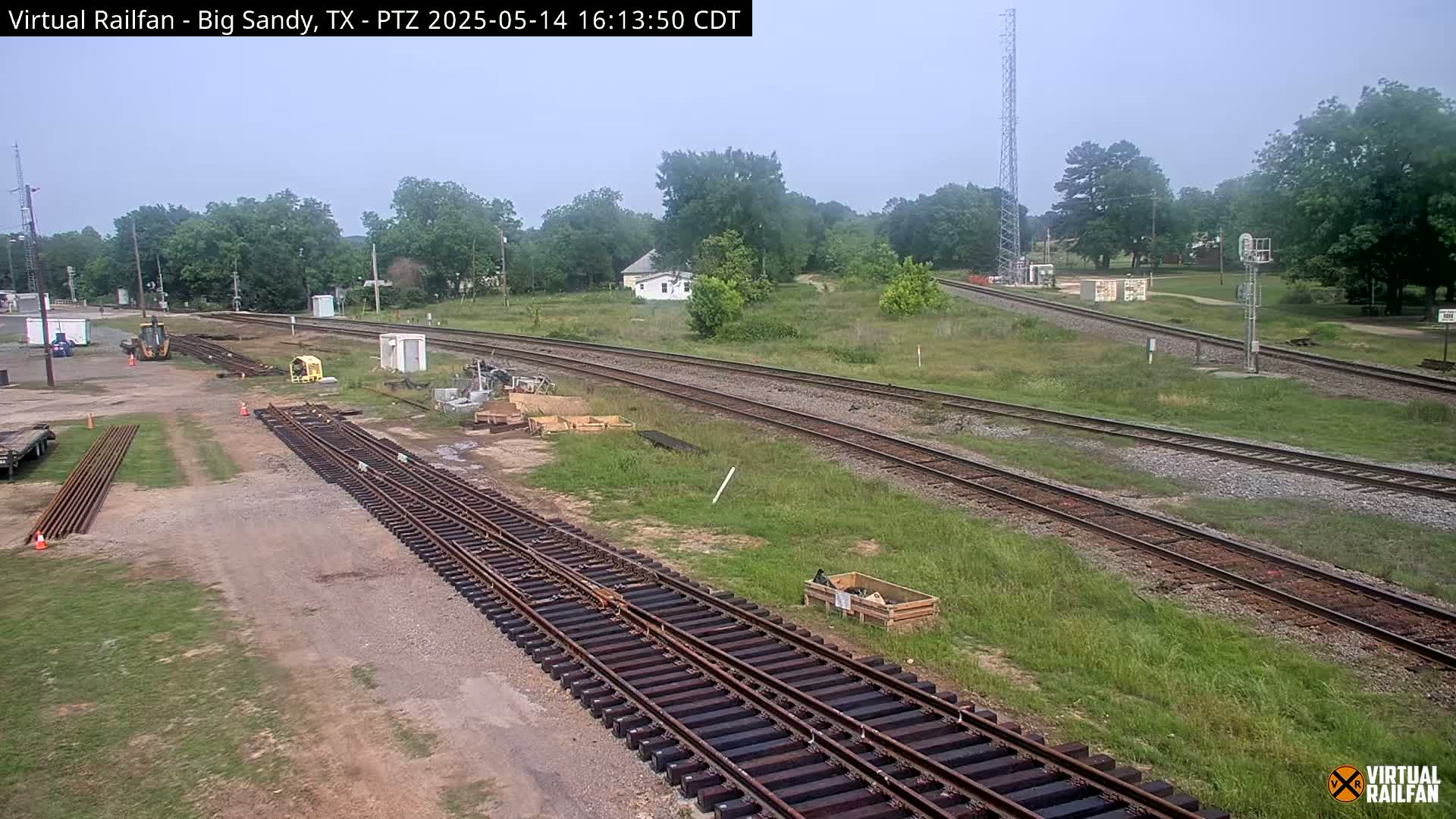 A mostly clear day shows railroad tracks under construction, with several pieces of heavy machinery and materials present near a grassy area with trees in the background.
