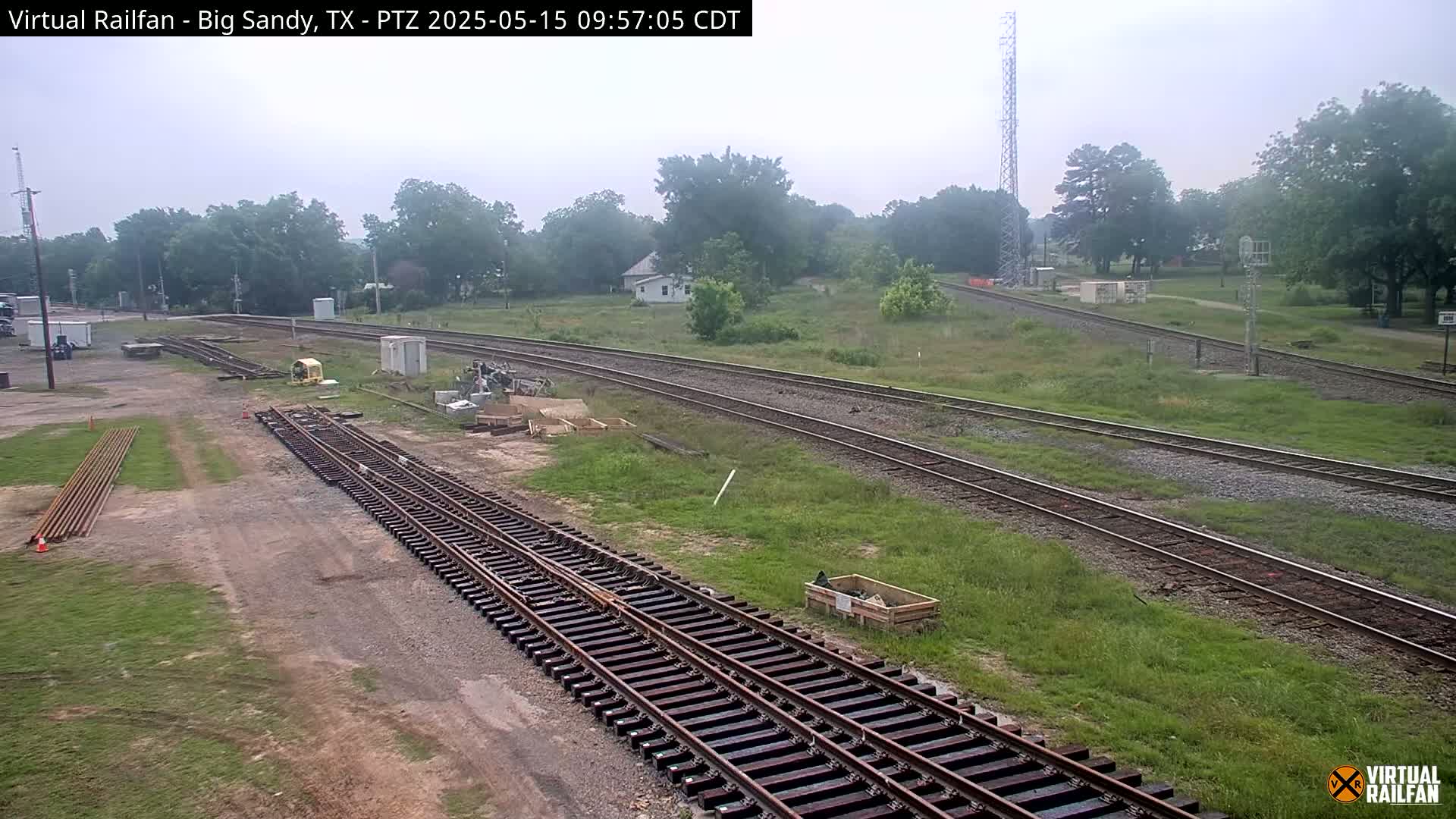 A hazy view of railroad tracks under construction, surrounded by overgrown grass and a few buildings in the distance.