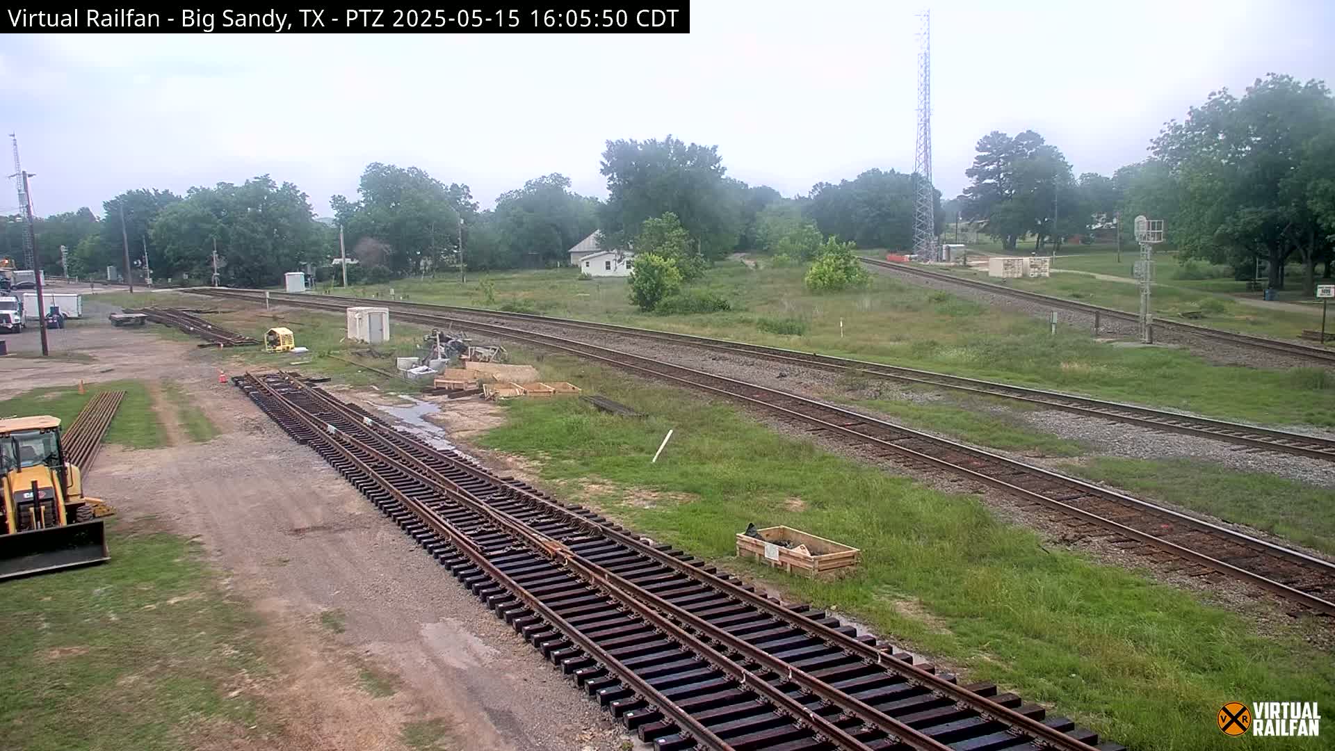 Railroad tracks undergoing construction or maintenance are situated in a grassy area with some buildings and trees visible in the background under an overcast sky.