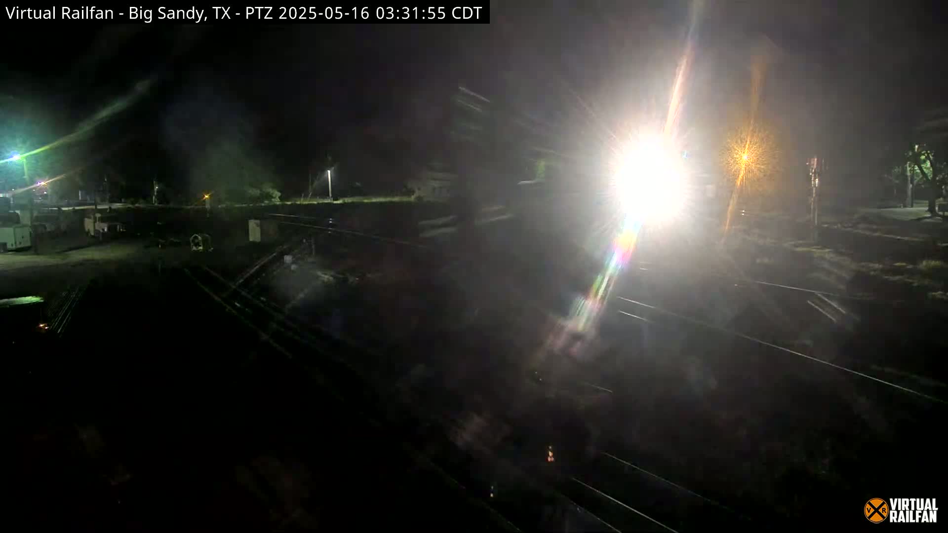 A nighttime view of a train's bright headlight illuminating railroad tracks and surrounding area.