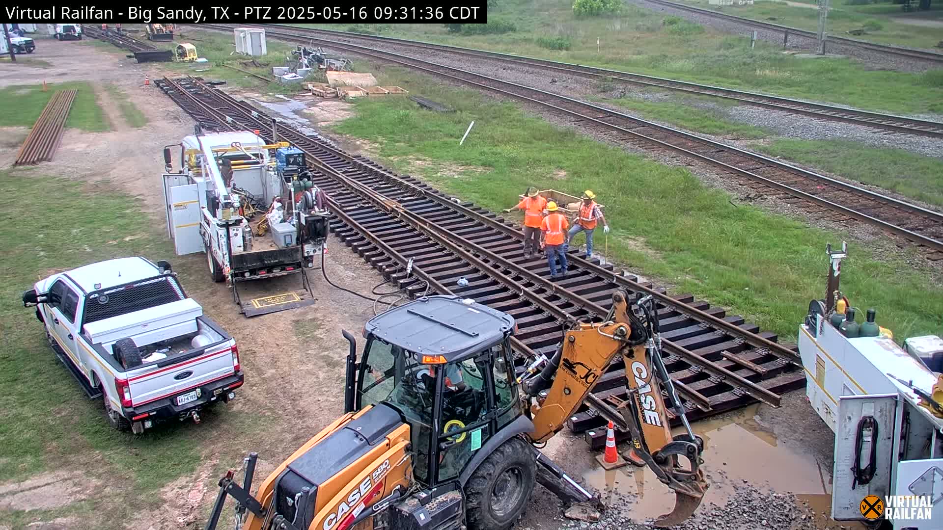 Construction workers are repairing railroad tracks with heavy machinery on a partly cloudy day.