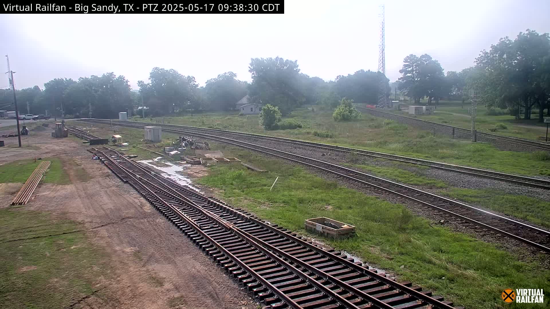 A hazy outdoor scene shows railroad tracks undergoing construction or maintenance, with some equipment present and greenery surrounding the tracks.