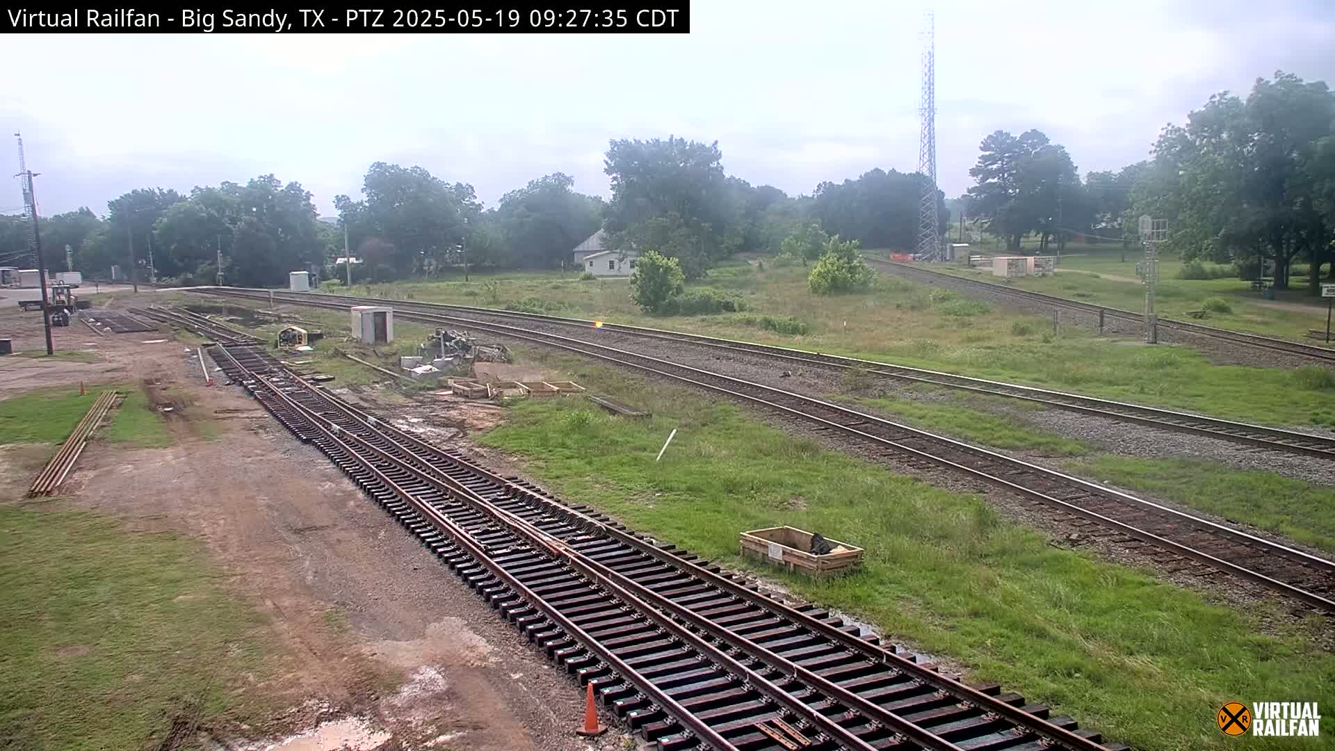 A view of railroad tracks under a partly cloudy sky, with some construction or maintenance work visible near the tracks and surrounding grassy areas.