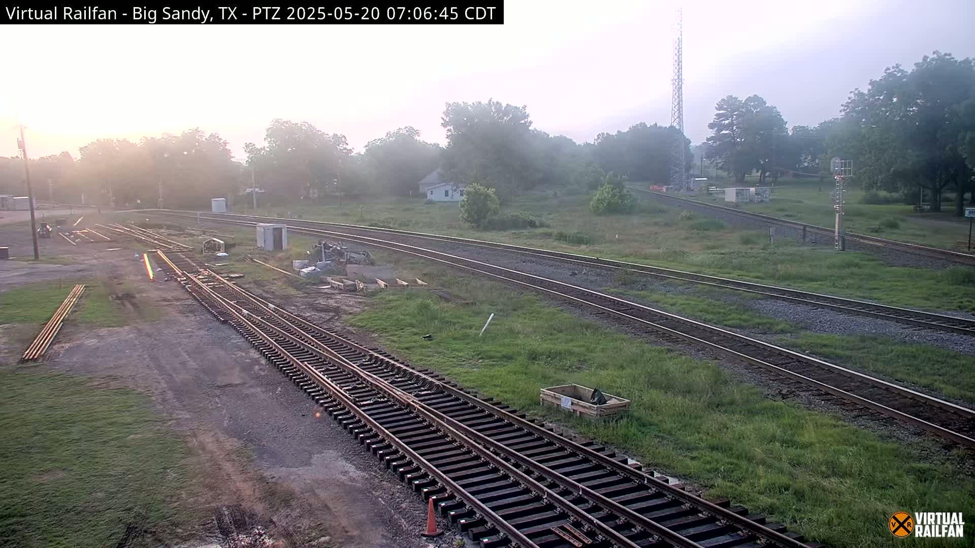 Several sets of train tracks wind through a grassy area with some light fog under a hazy sunrise.