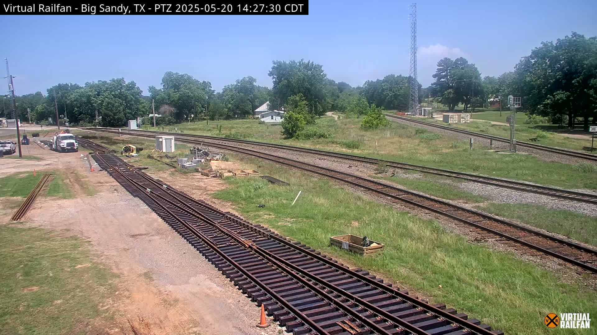 A sunny day reveals railroad tracks undergoing construction, with new track sections laid near a truck and some construction equipment, set against a backdrop of trees and grassy fields.