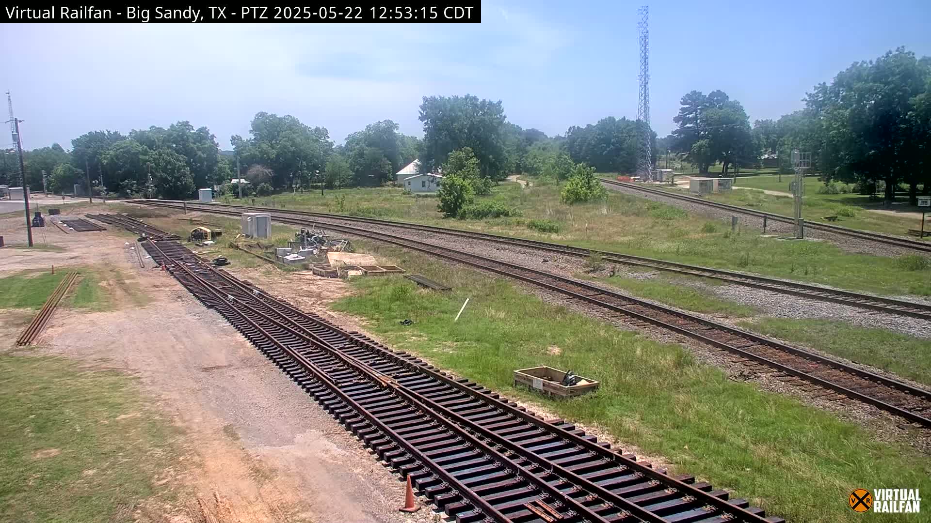 Under a sunny sky, railroad tracks snake through a grassy area with some construction equipment and a small building visible in the distance.