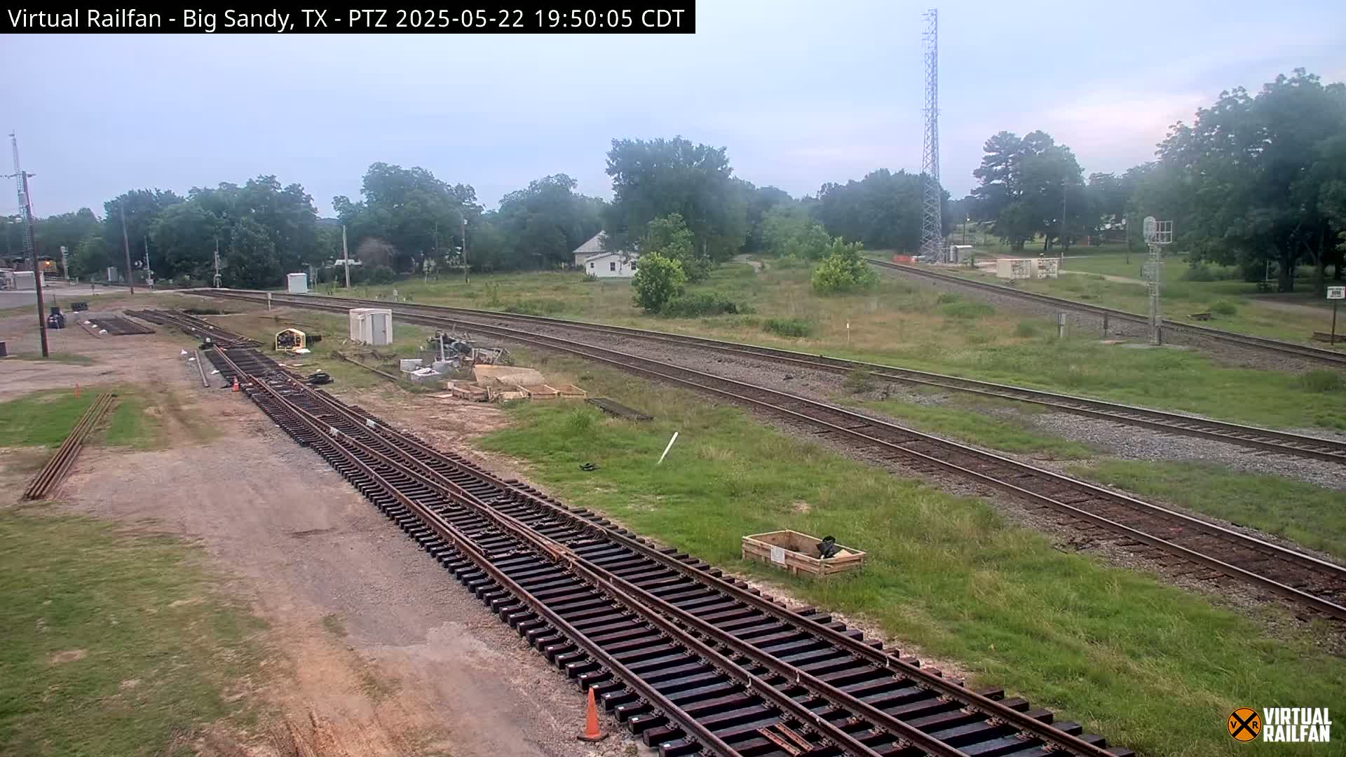 Under an overcast sky, railroad tracks undergoing construction are shown next to overgrown grassy areas and a few buildings.