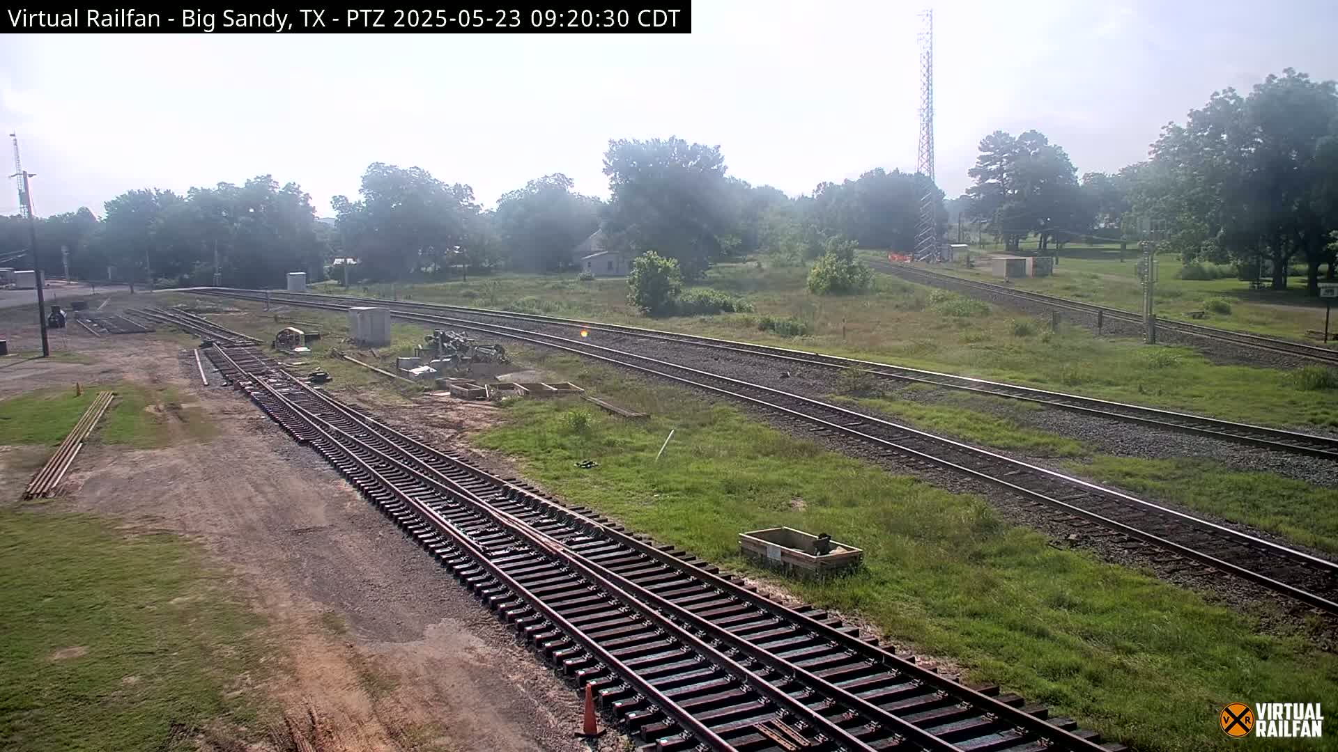 Several sets of train tracks weave through a grassy area under a hazy sky, with construction or maintenance work visible near some of the tracks.