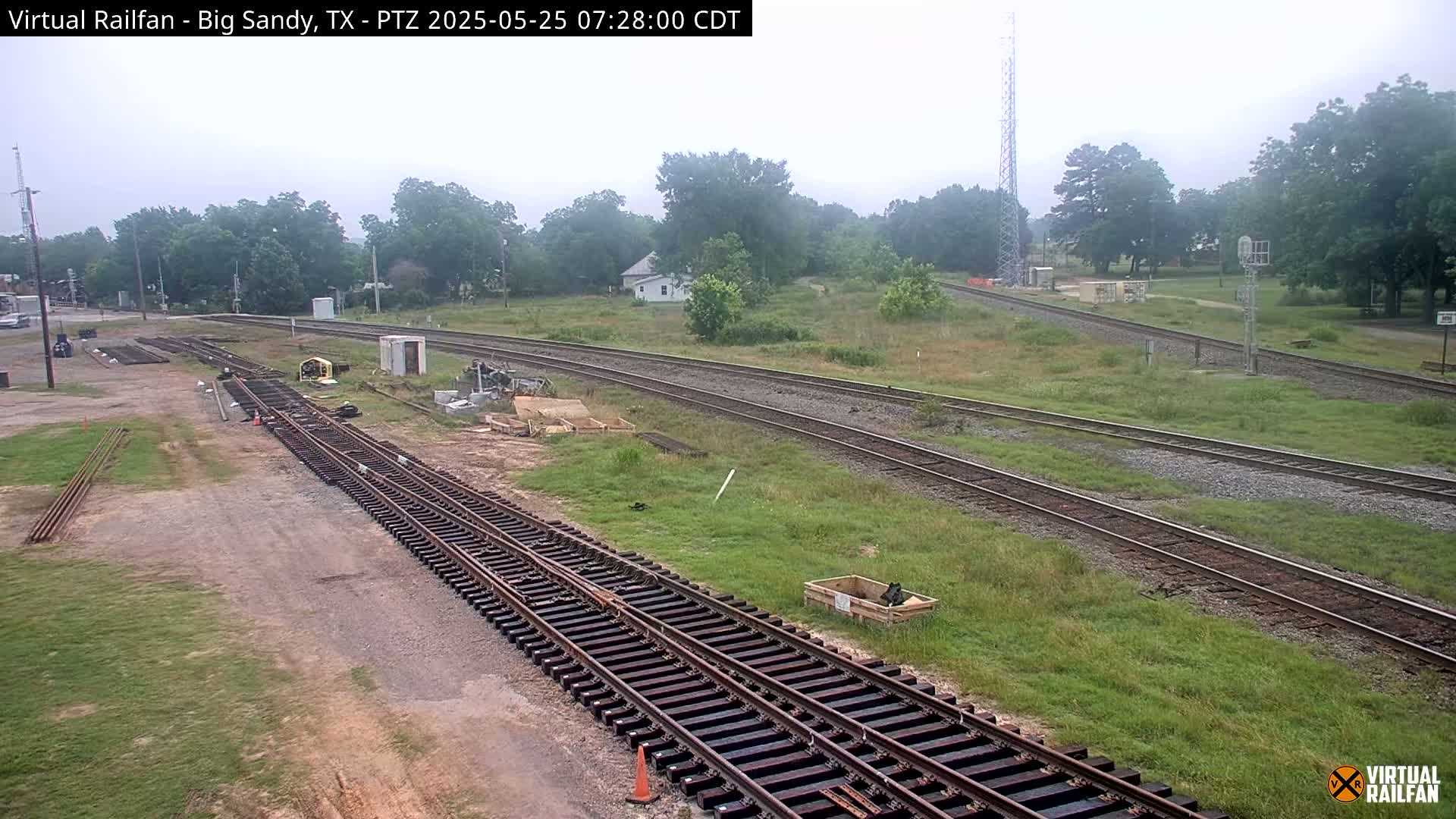 Railroad tracks under construction are visible, alongside existing tracks that curve through a grassy area with trees and a single house in the background, all under an overcast sky.