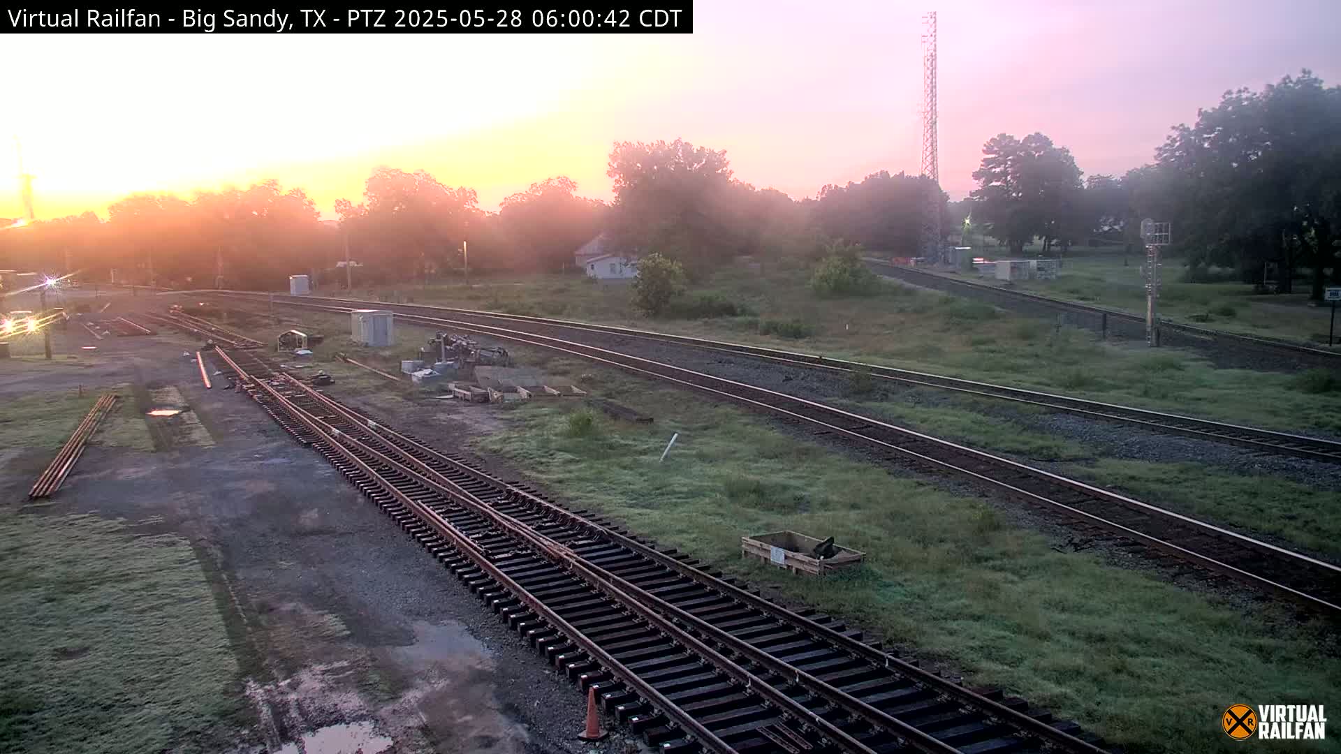 Several sets of railroad tracks intersect near a grassy area at sunrise, with a light fog hanging in the air.