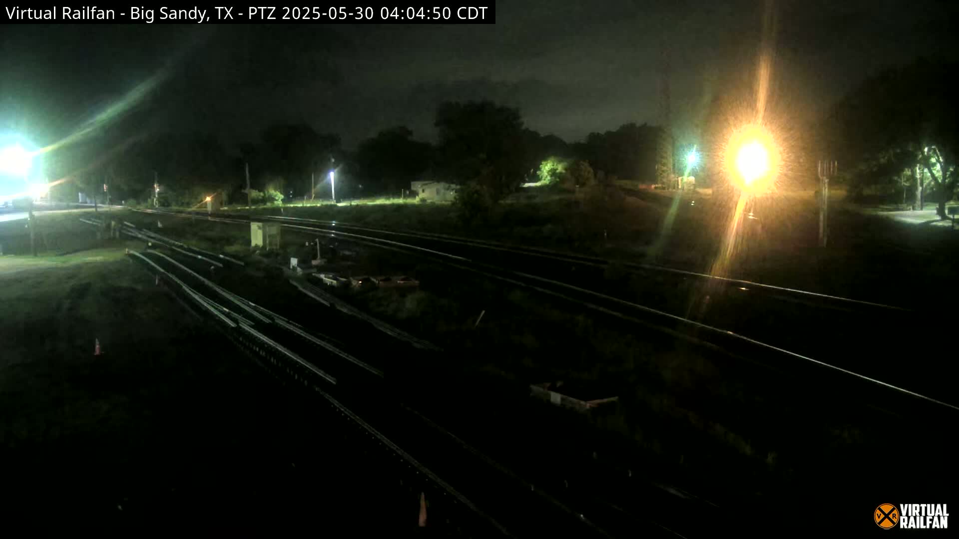 A nighttime view of several sets of train tracks, surrounded by dark vegetation under a cloudy sky.