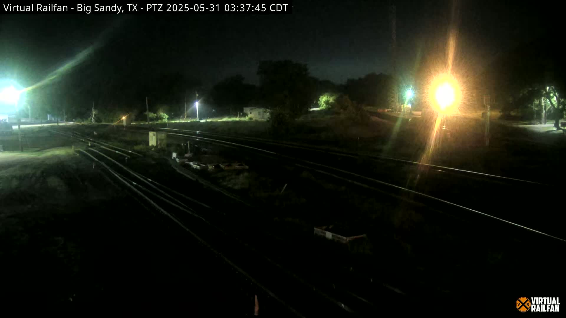 At night, a high-angle, long shot shows several sets of train tracks cutting across a dark, mostly empty area, with some vegetation and light sources visible in the distance.