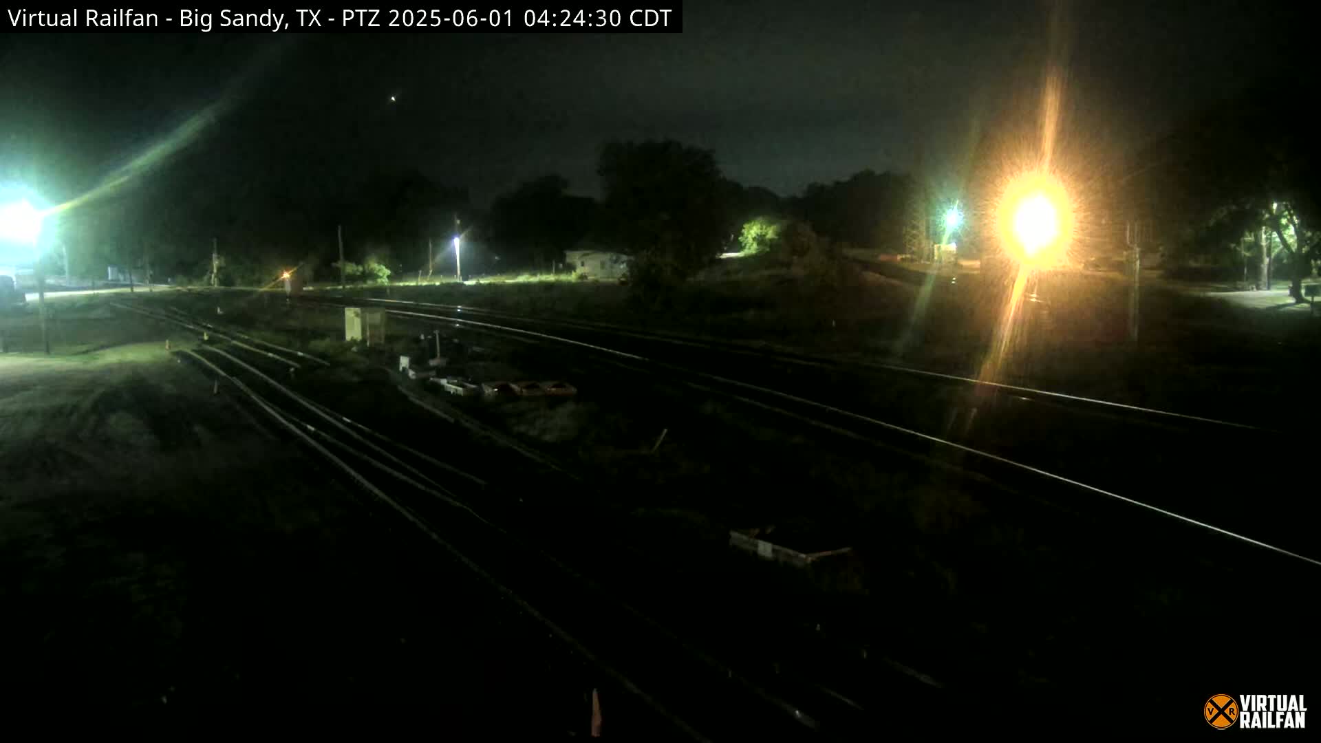 A nighttime, outdoor view of several railroad tracks under partly cloudy skies, with some vegetation and distant lights visible.
