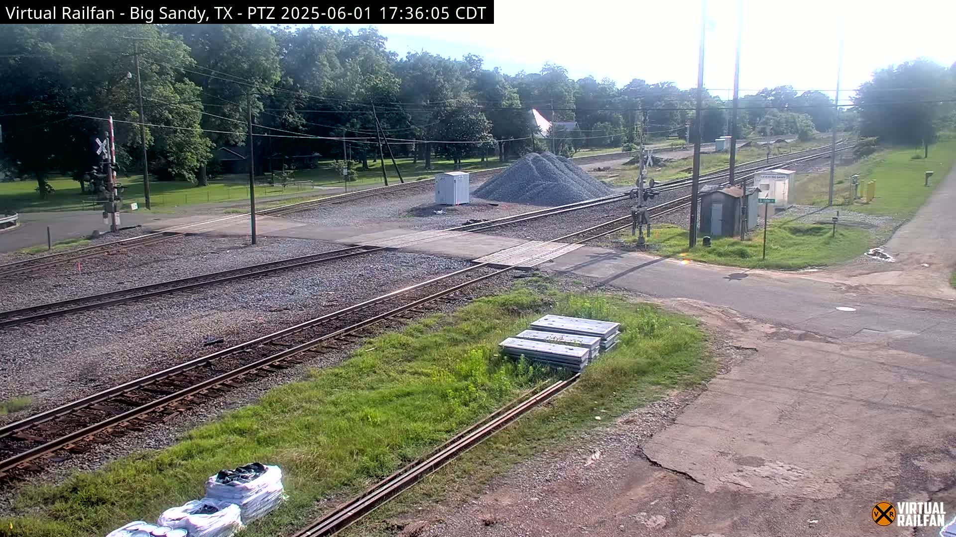 A railroad crossing with multiple sets of tracks, a pile of gravel, and some construction materials is visible under a partly cloudy sky.