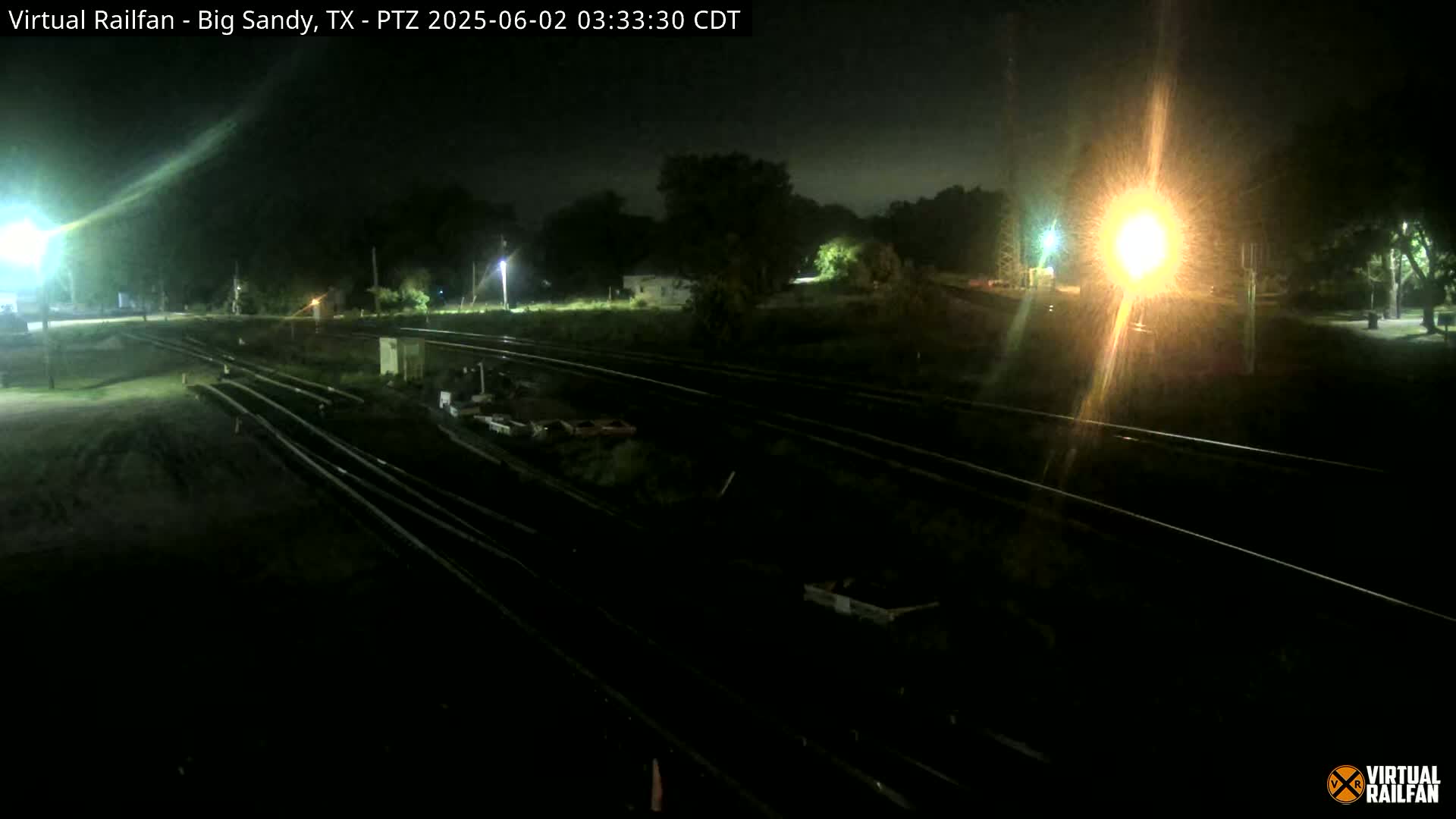 At night, under a dark sky, several sets of train tracks are visible, illuminated by artificial lights, with some vegetation and structures in the background.
