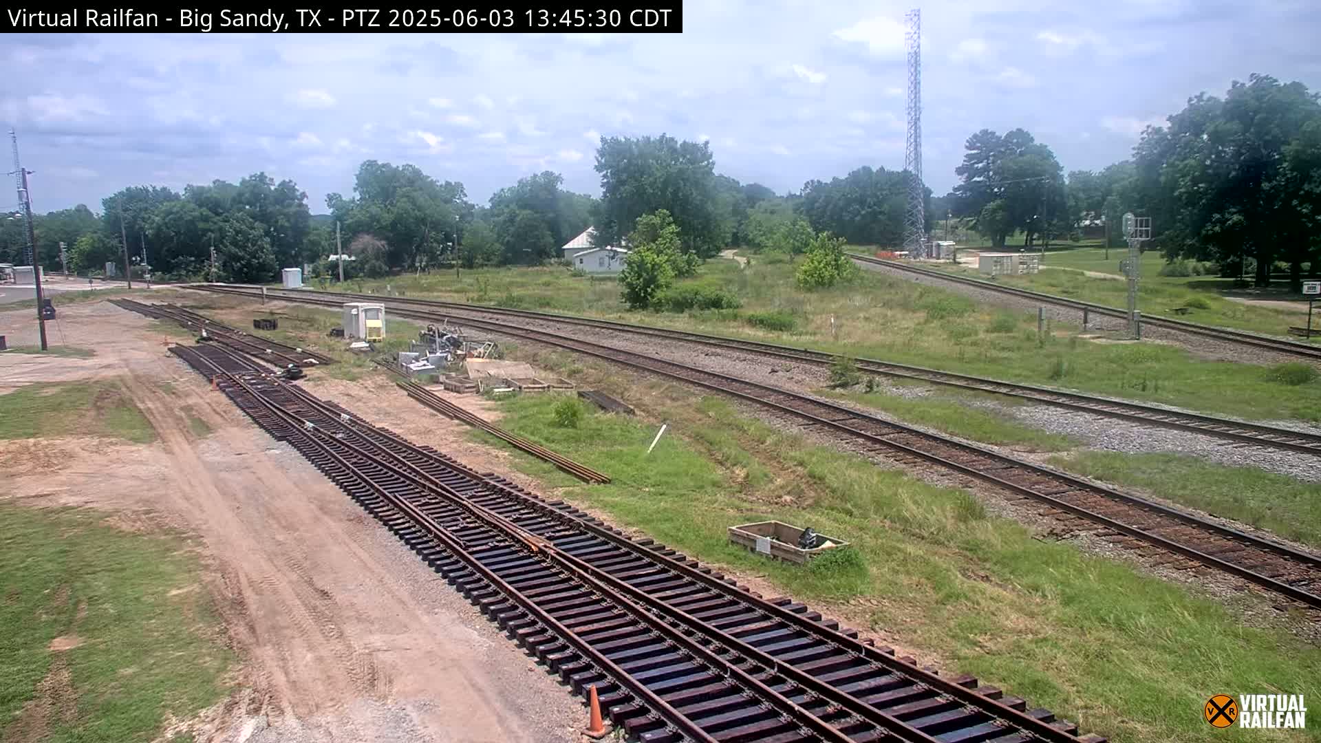 Under a partly cloudy sky, several sets of train tracks snake through a grassy area, with some construction or maintenance equipment visible near the tracks.