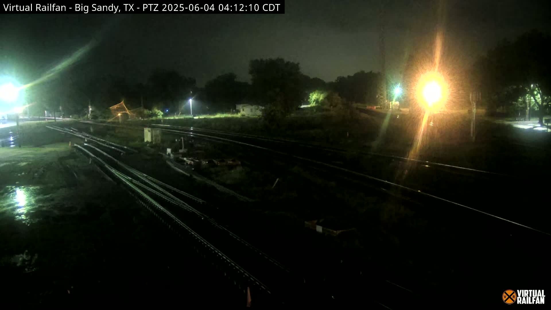 A nighttime view of rain-slicked railroad tracks and surrounding vegetation under a dark sky illuminated by artificial lights.
