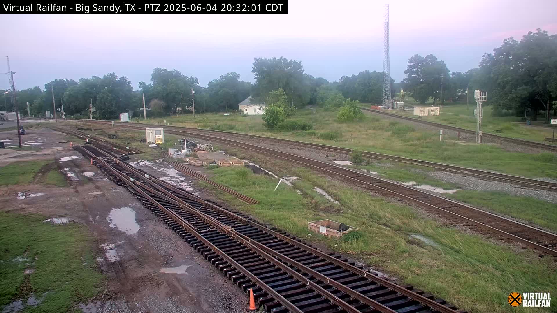 A railroad yard with multiple sets of tracks, some undergoing construction or maintenance, sits in a grassy area under a twilight sky.