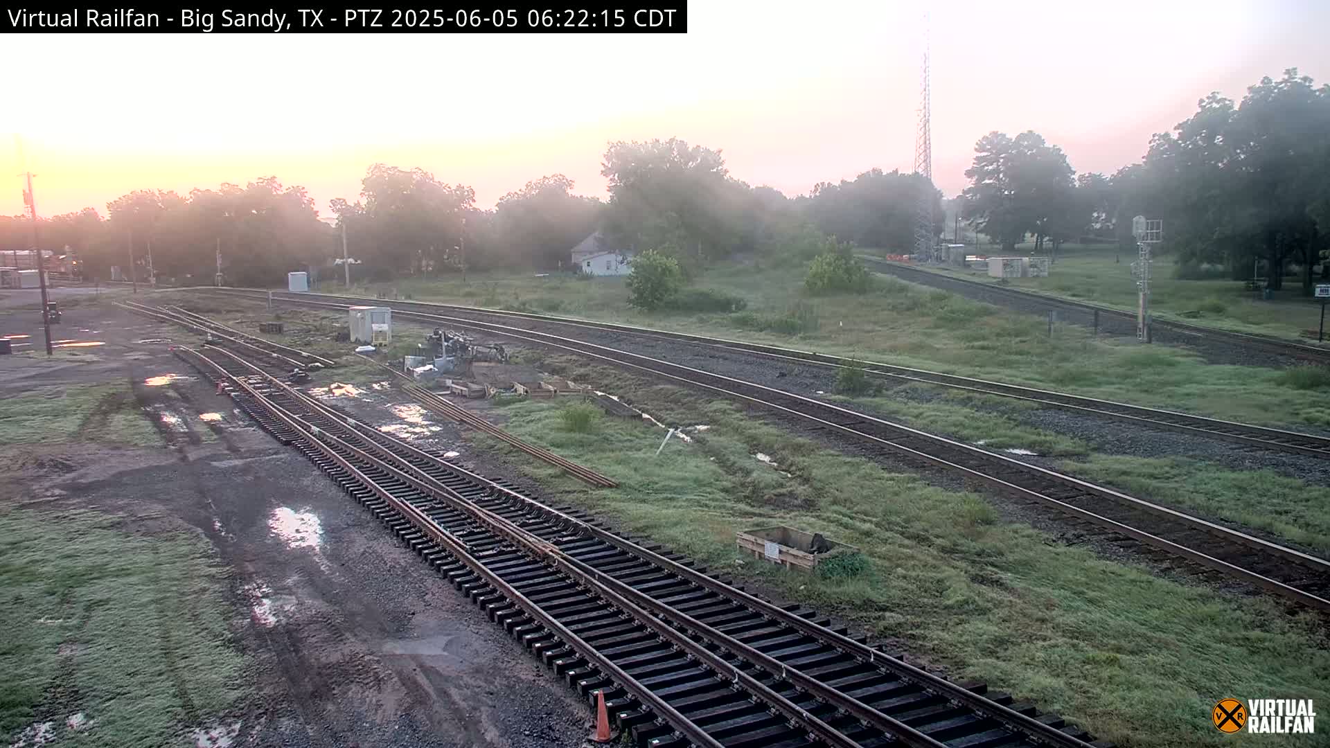 Several sets of train tracks curve through a grassy area at sunrise, with some puddles present on the ground.