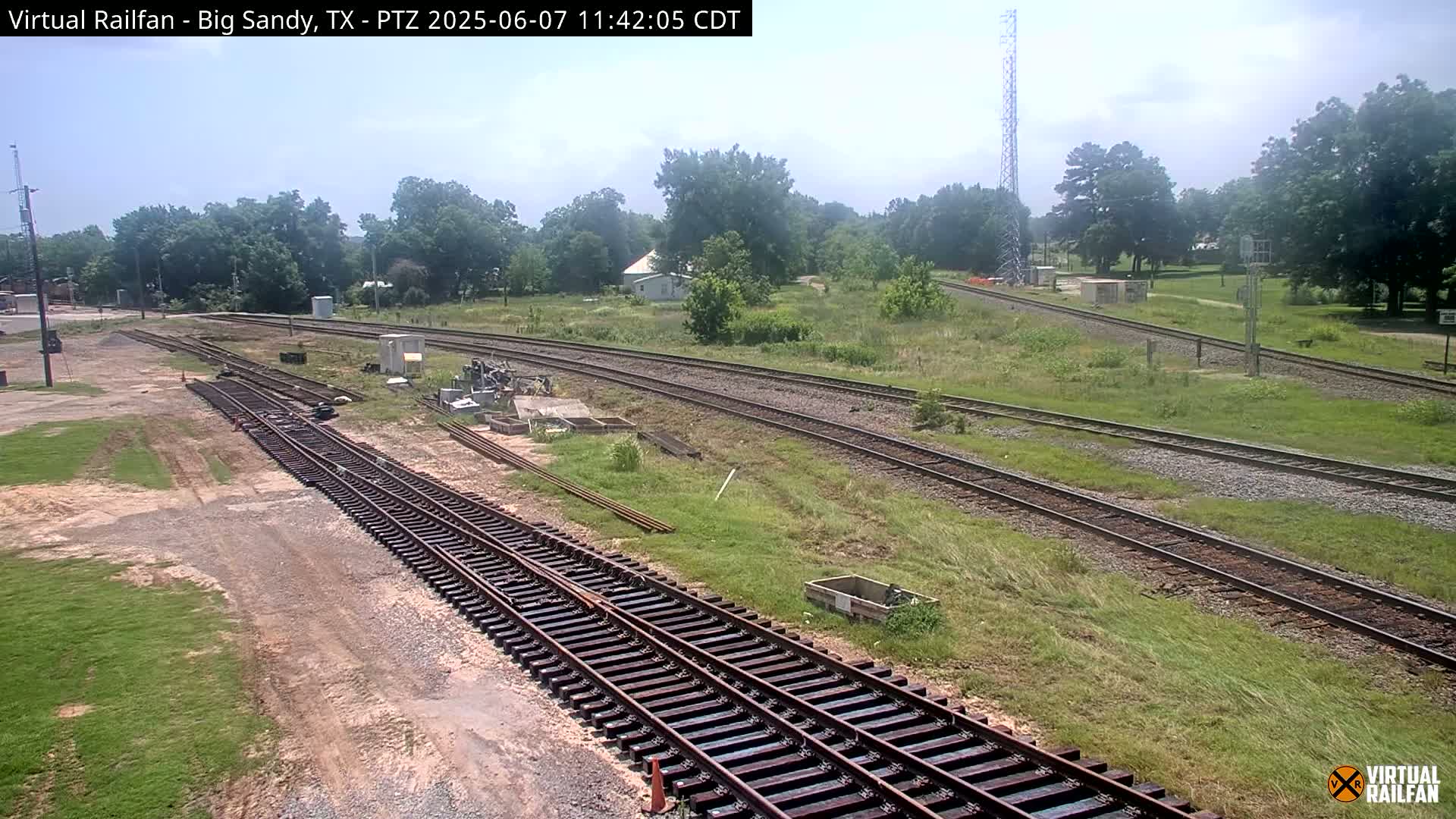 A sunny day view of railroad tracks and switching yards surrounded by green grass and trees.