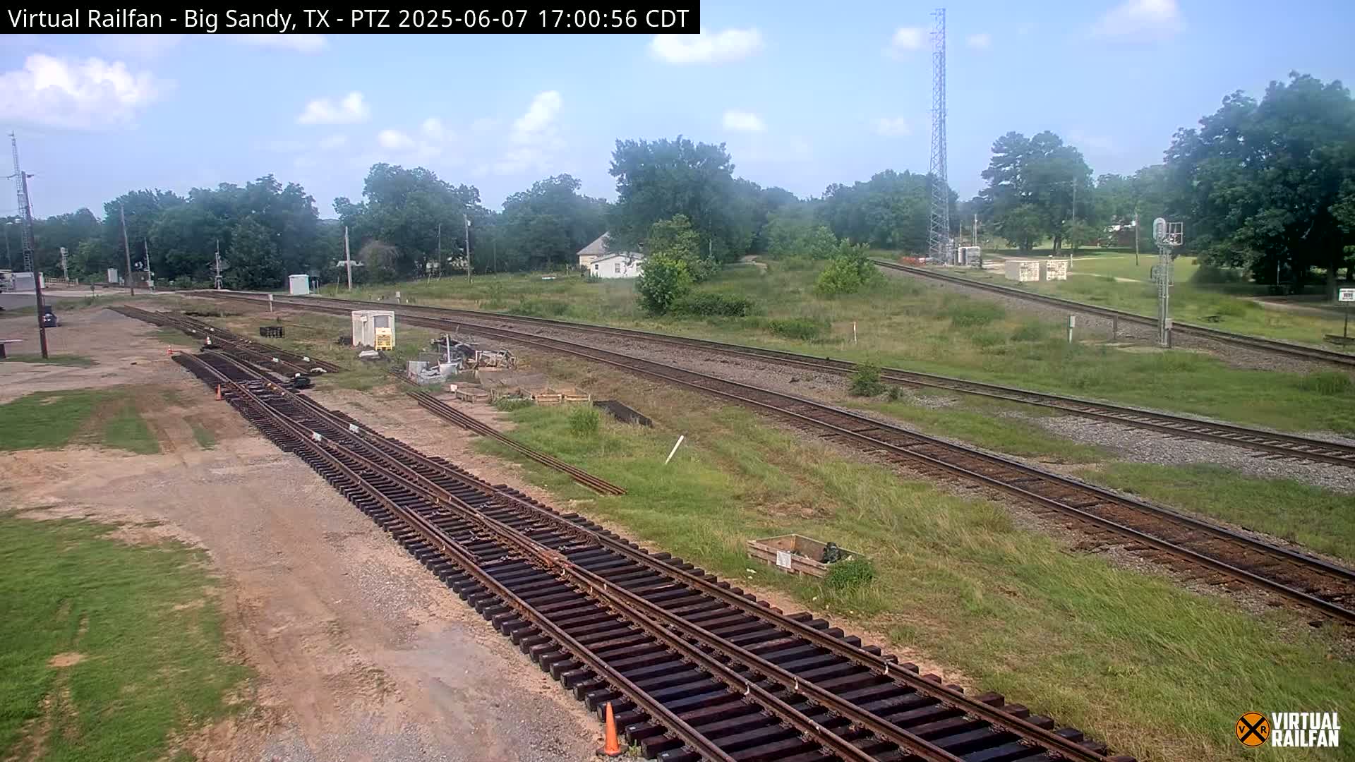 Several sets of train tracks wind through a grassy area under a partly cloudy sky, with some trees and small buildings visible in the background.