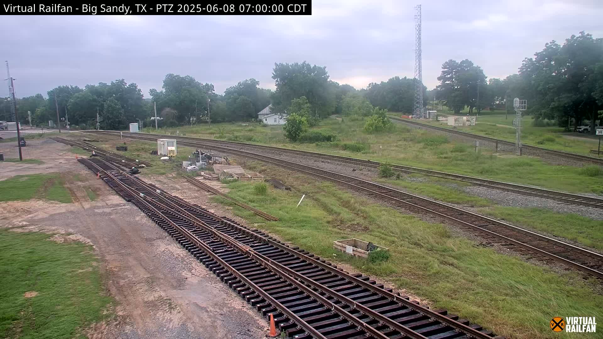 A view of railroad tracks undergoing construction or maintenance, under an overcast sky, in a rural setting with some trees and buildings in the distance.