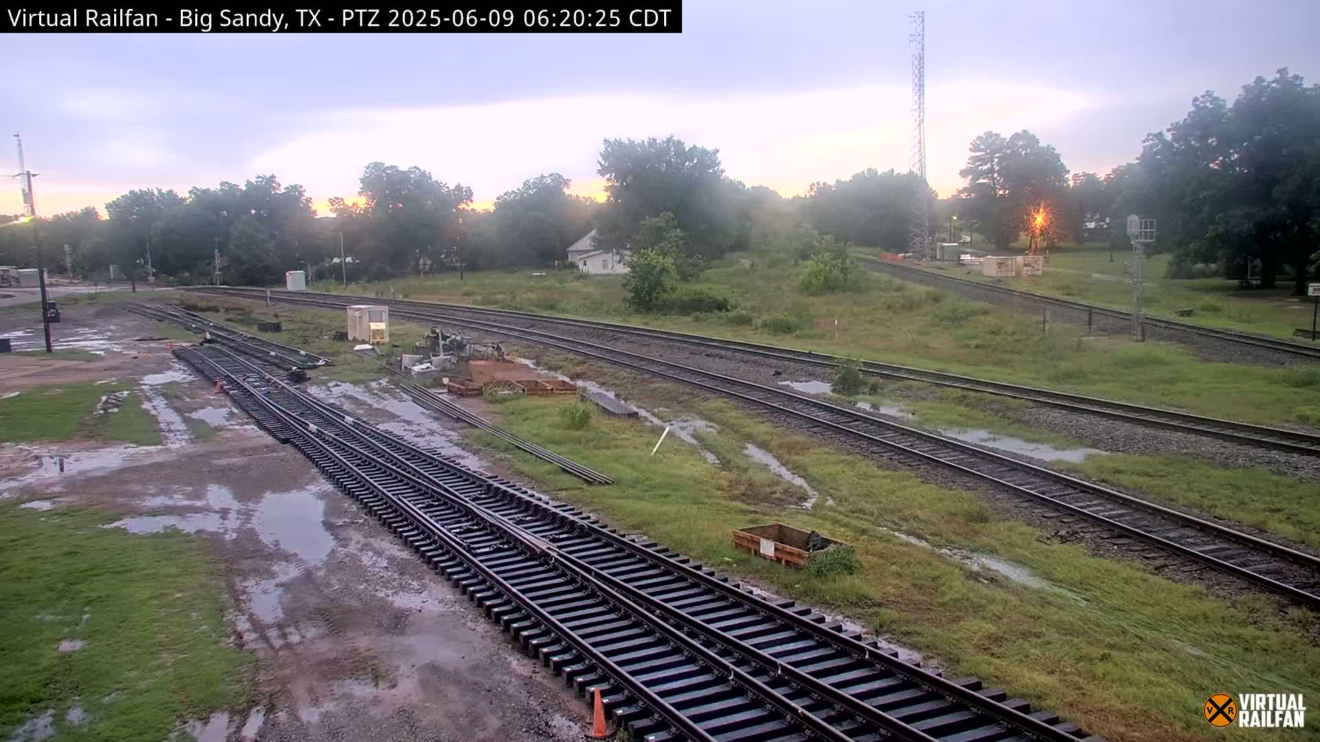 Several sets of railroad tracks wind through a grassy area with puddles of water, under a partly cloudy sky at sunrise.