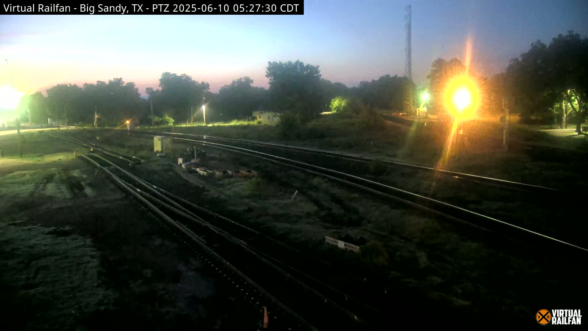 Several sets of train tracks are visible at twilight, surrounded by overgrown vegetation and illuminated by artificial lights.