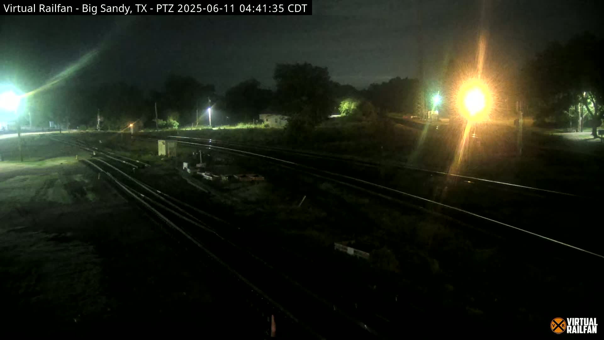 A nighttime view of railroad tracks and surrounding vegetation, illuminated by artificial lights under a dark sky.
