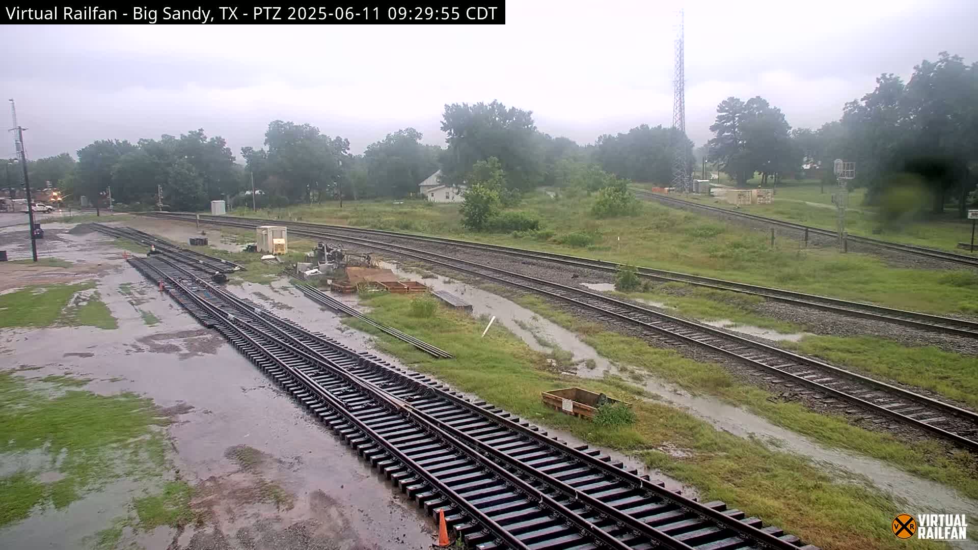 Several sets of railroad tracks are partially submerged in standing water during a rainy day, surrounded by grassy areas and trees.