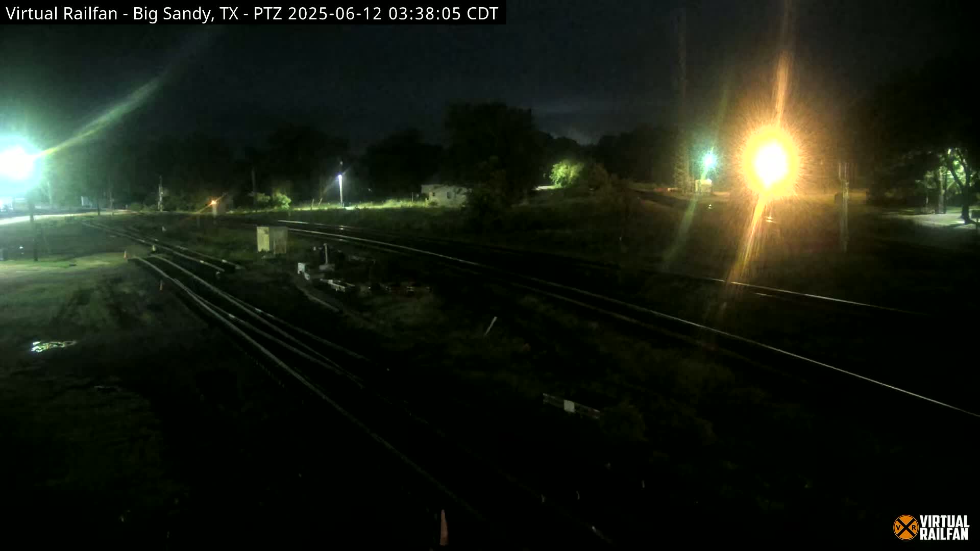 At night, several sets of train tracks are visible, surrounded by dark, overgrown vegetation under artificial light.
