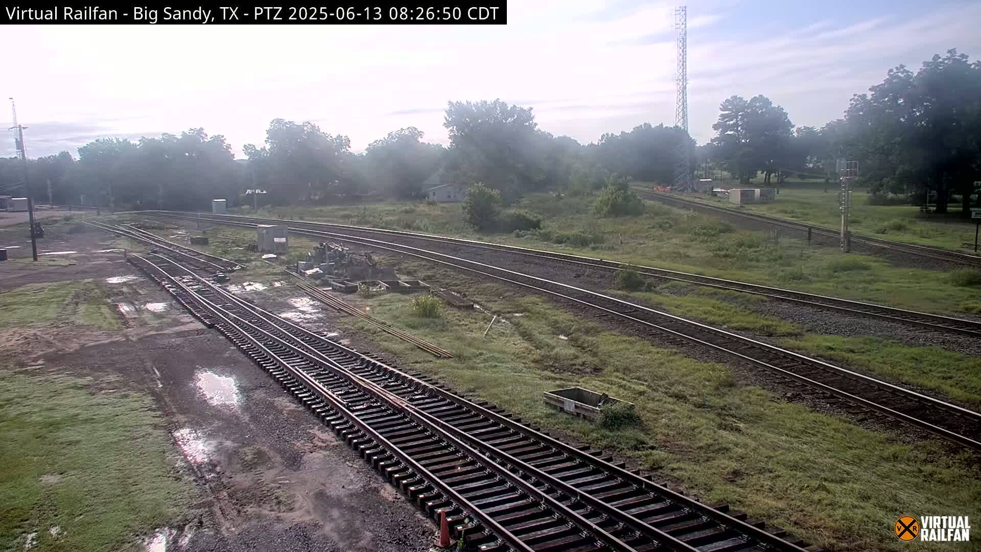 Several sets of train tracks curve through a grassy area with some puddles under a hazy sky.