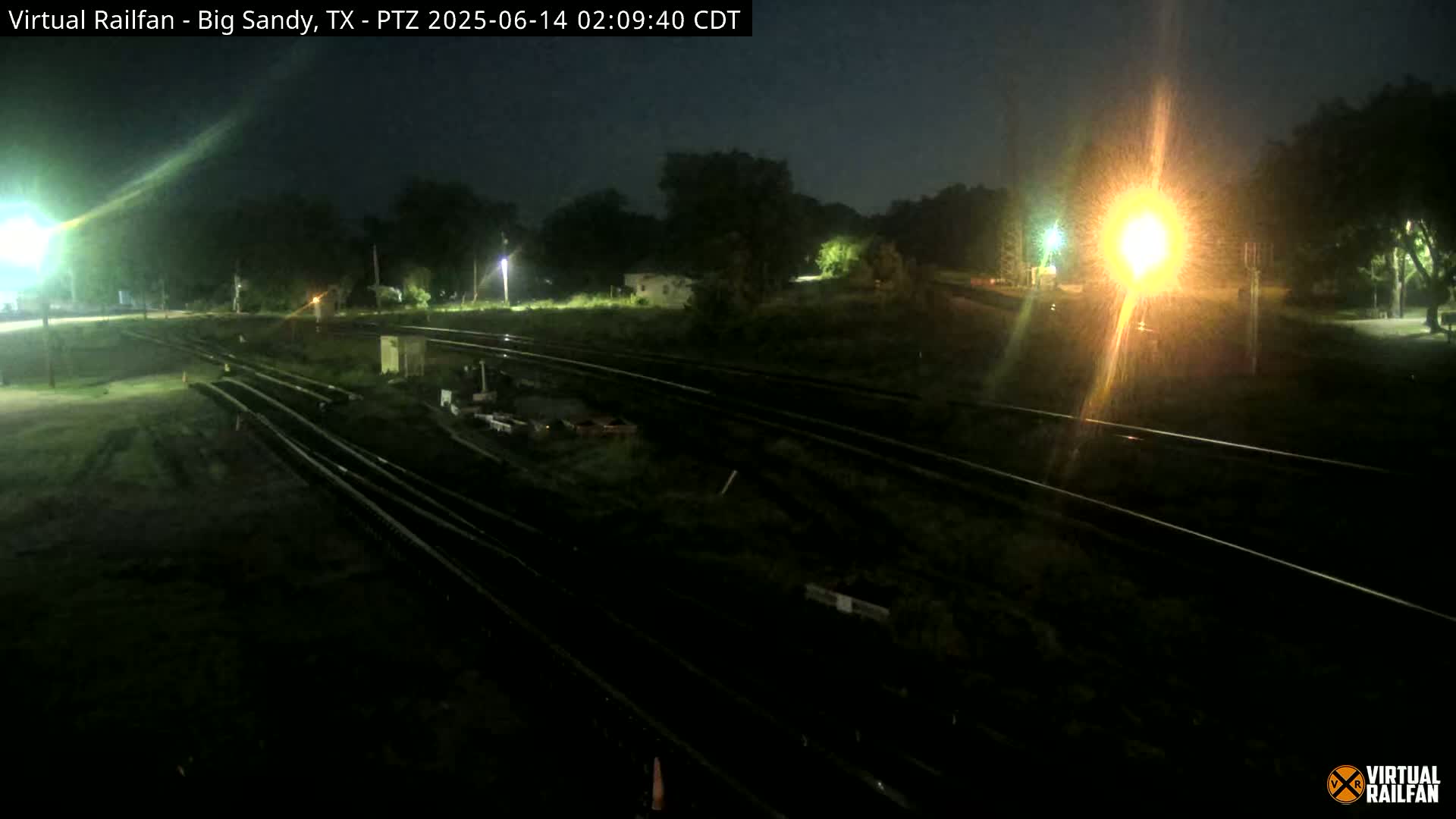 A nighttime, high-angle view of several sets of train tracks surrounded by dark vegetation and illuminated by artificial lights.