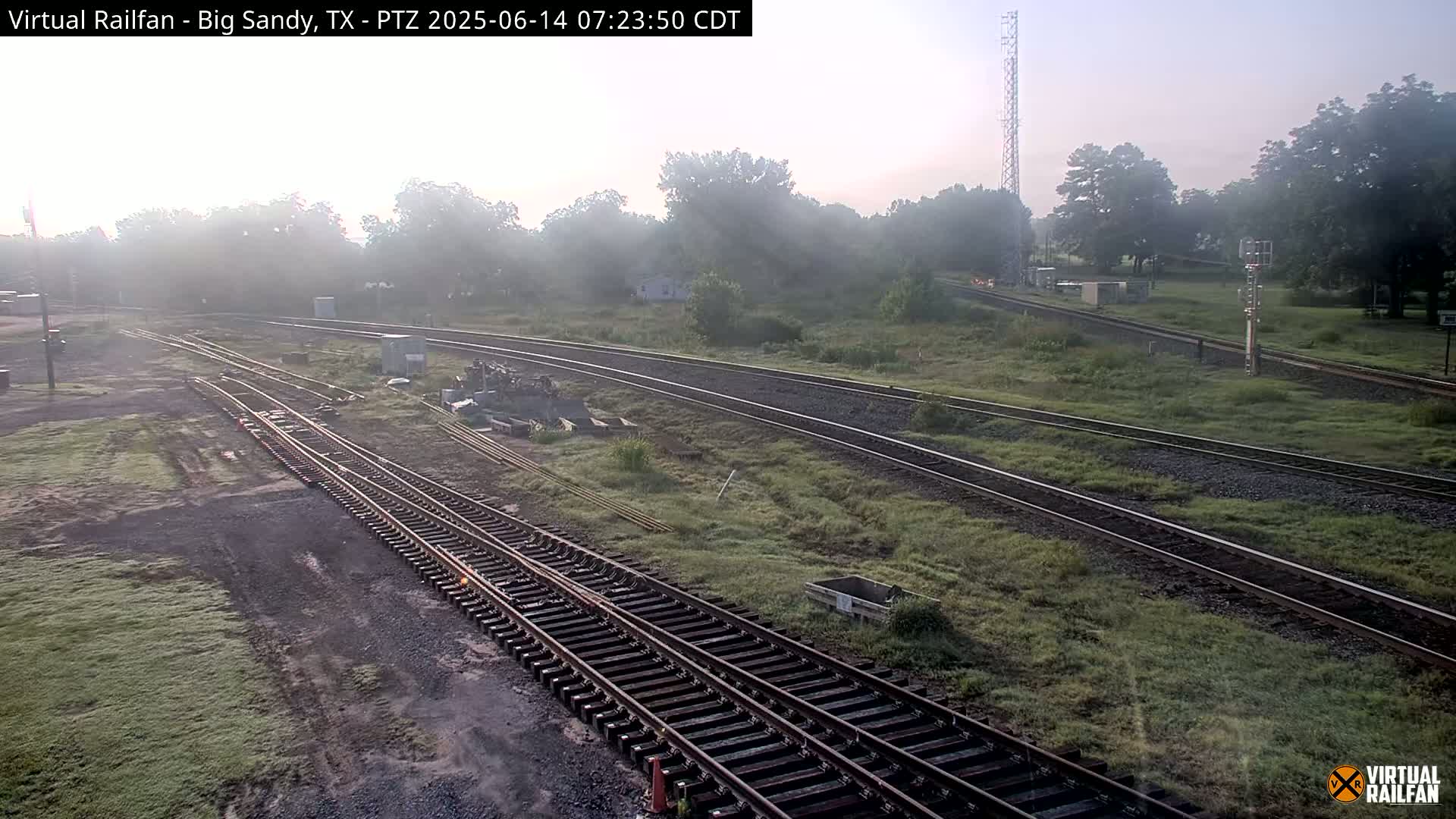 A hazy morning view of multiple sets of train tracks that curve around grassy areas and trees.