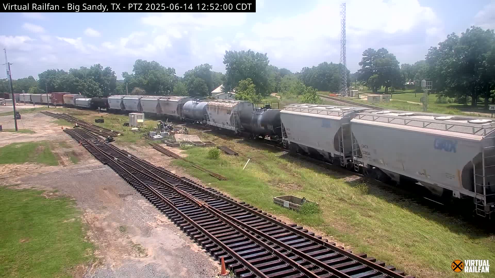 A long freight train sits on railroad tracks under a partly cloudy sky, surrounded by green grass and trees.