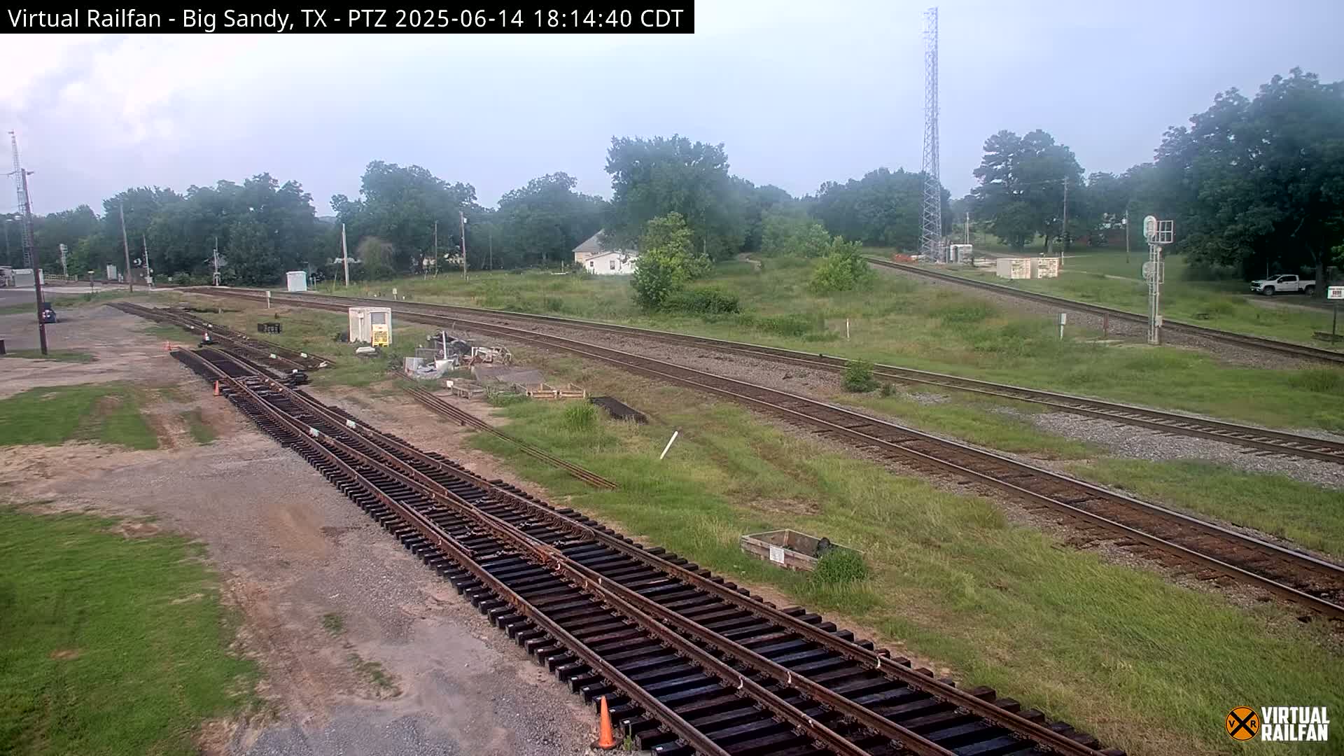 A long freight train sits on railroad tracks under a partly cloudy sky, surrounded by green grass and trees.