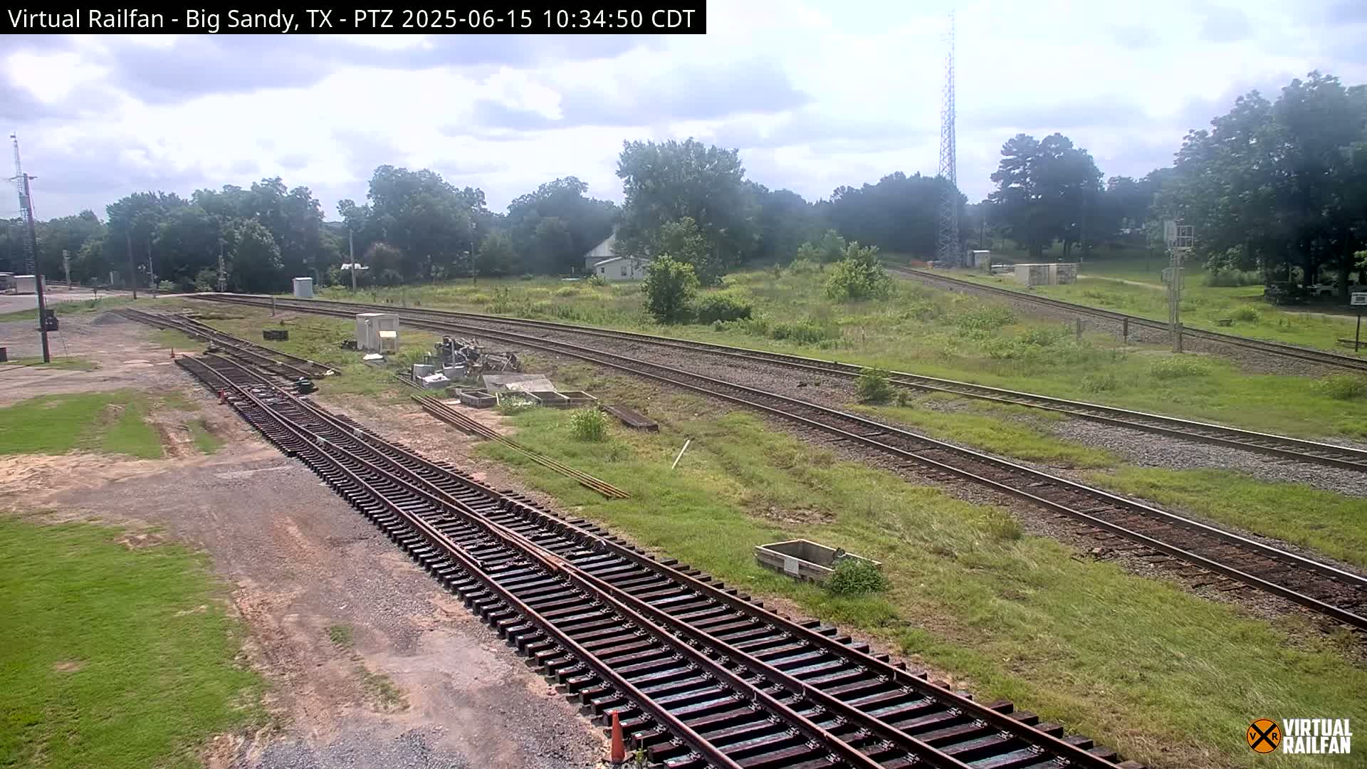 Under a partly cloudy sky, several sets of train tracks wind through a grassy area with some trees and small buildings in the background.