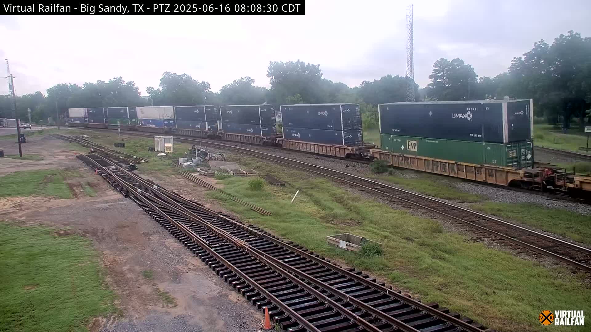 A long freight train carrying multiple shipping containers moves along a railroad track on an overcast day.