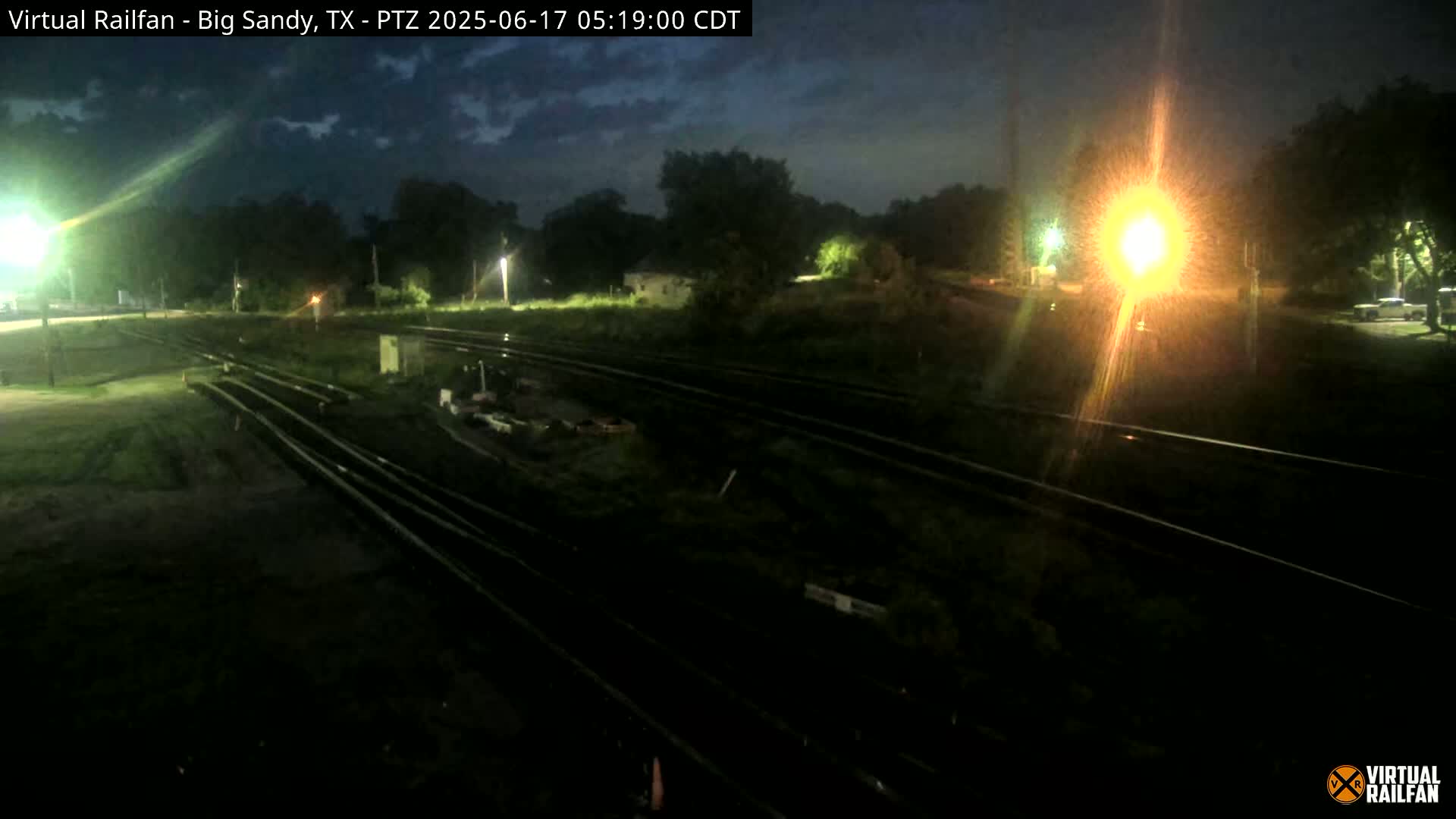 A nighttime view of train tracks in a dimly lit area with some vegetation and a few lights visible.