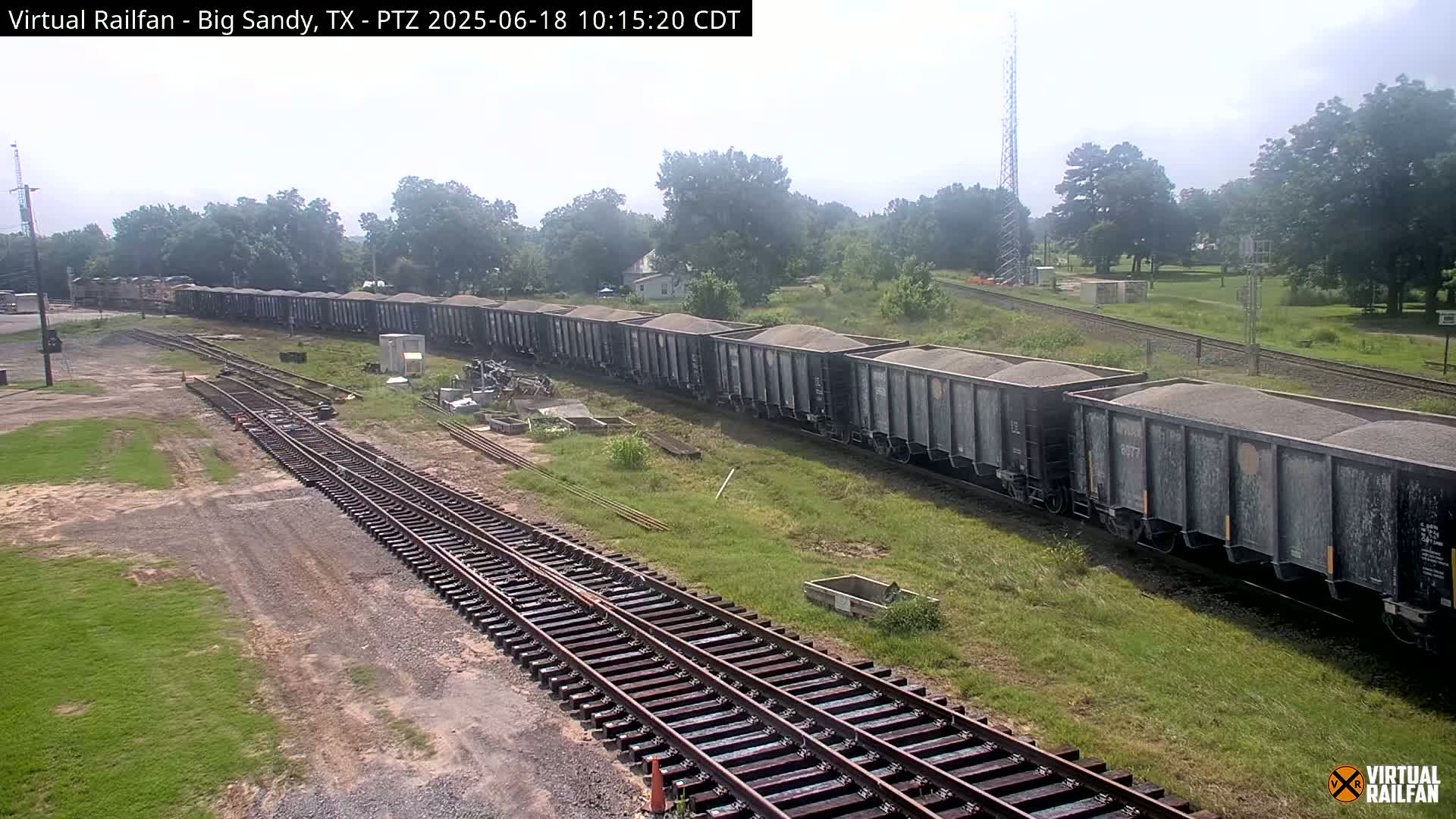 A long freight train carrying a load of what appears to be dark-colored gravel sits on a railway track on a partly sunny day.