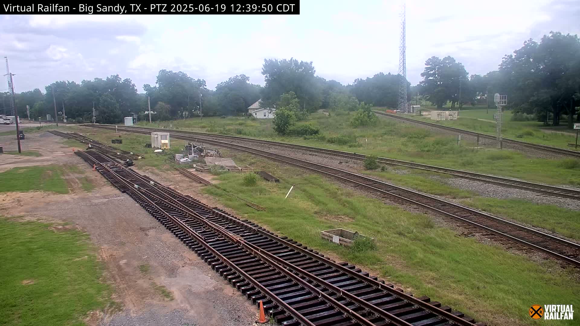 A sunny day reveals several sets of train tracks winding through a grassy area, with a small building and trees in the background.