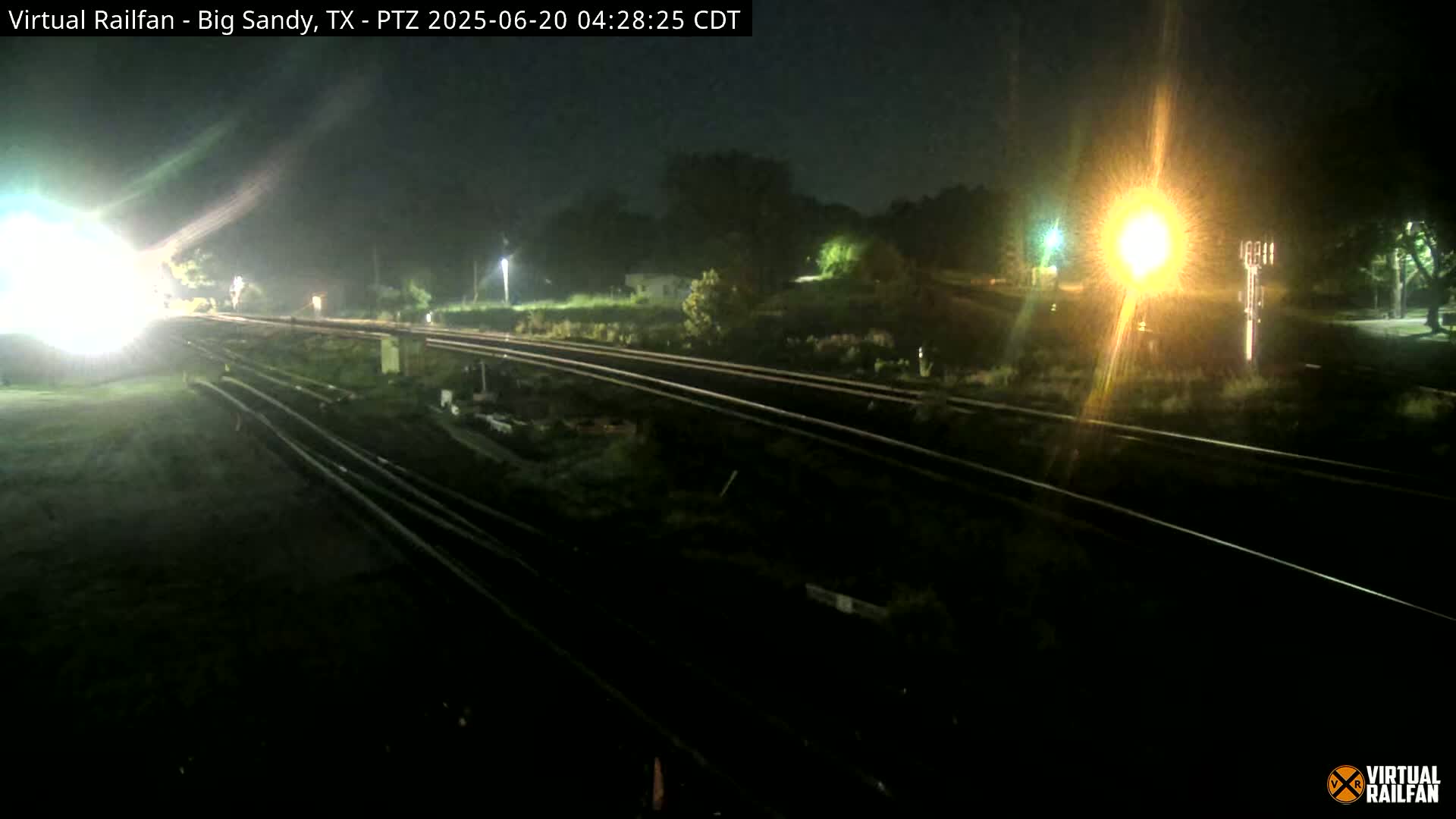 At night, several sets of train tracks are visible, illuminated by bright lights, with some greenery and trees in the background.